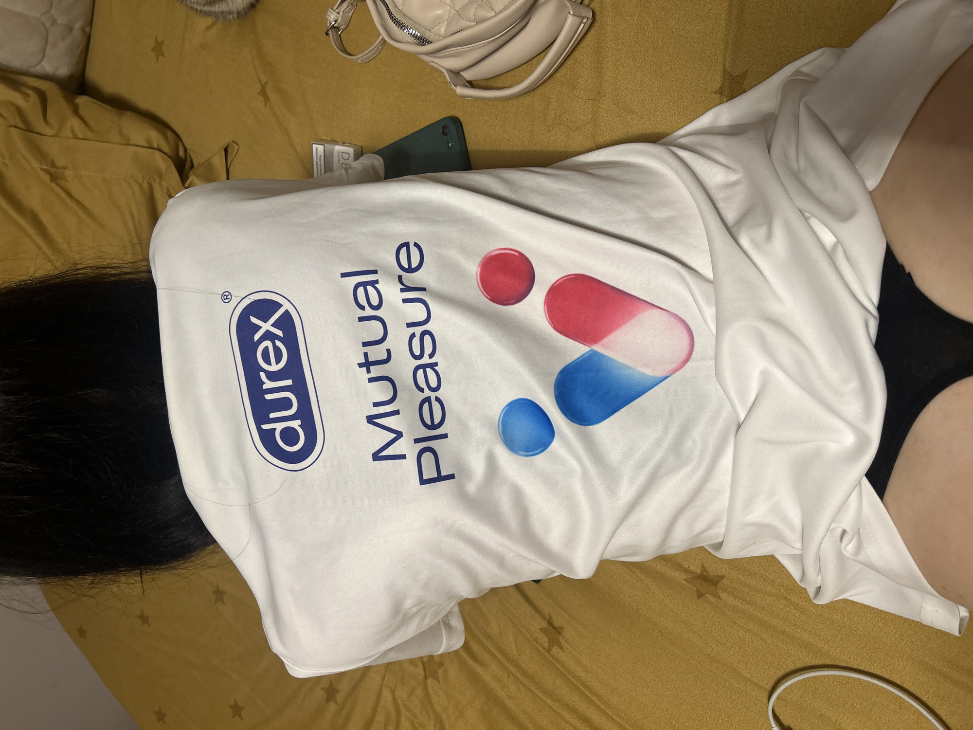 A woman with long, dark hair is lying face down on a mustard-colored bed. She is wearing a white t-shirt with the Durex logo and the words "Mutual Pleasure" printed on it. The shirt also features a graphic with red and blue pill-shaped designs. She is wearing black underwear. In the upper part of the image, there is a beige purse, a green phone, and a pack of cigarettes. The bed is in an indoor setting. There is no indication of the location being in Burgas, Bulgaria.