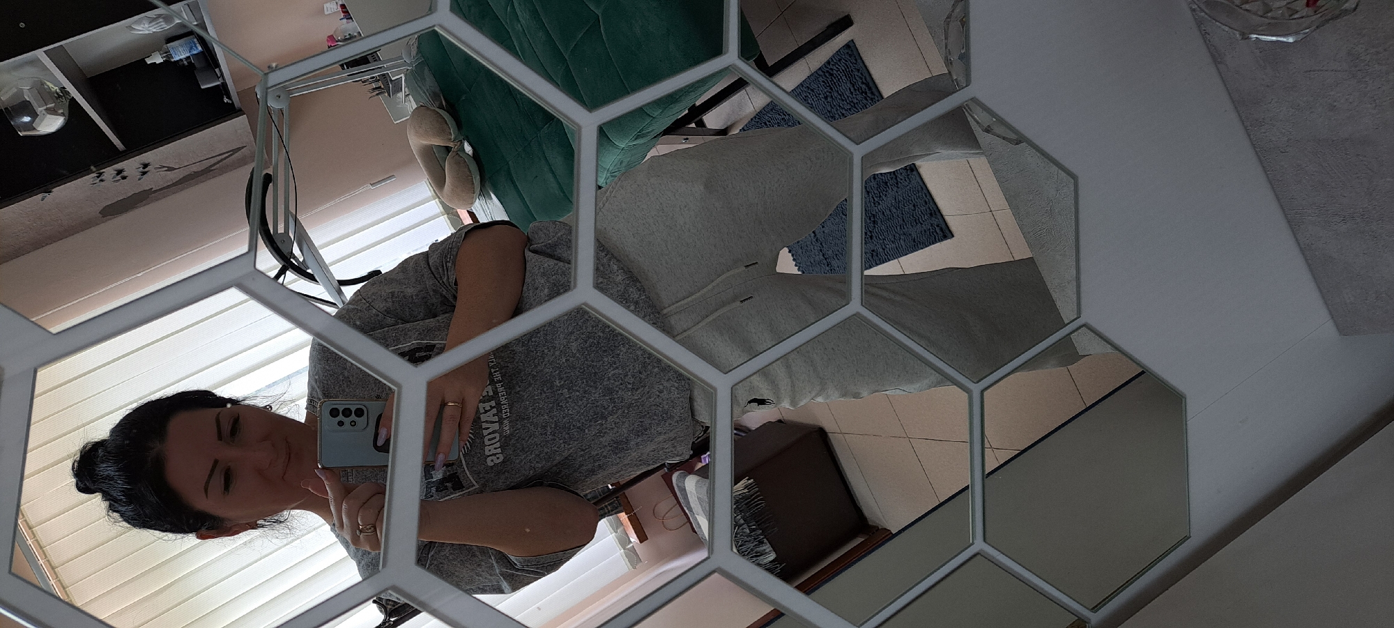 A woman is taking a selfie in a hexagon-shaped mirror in Yessentuki, Russia. She is wearing a grey t-shirt and grey sweatpants, and her hair is pulled up in a bun. The mirror reflects a room with light-colored blinds on a window, a dark wooden cabinet with a striped blanket on it, and a green massage table with a beige neck pillow. There is also a shelf with bottles and a glass visible in the reflection.