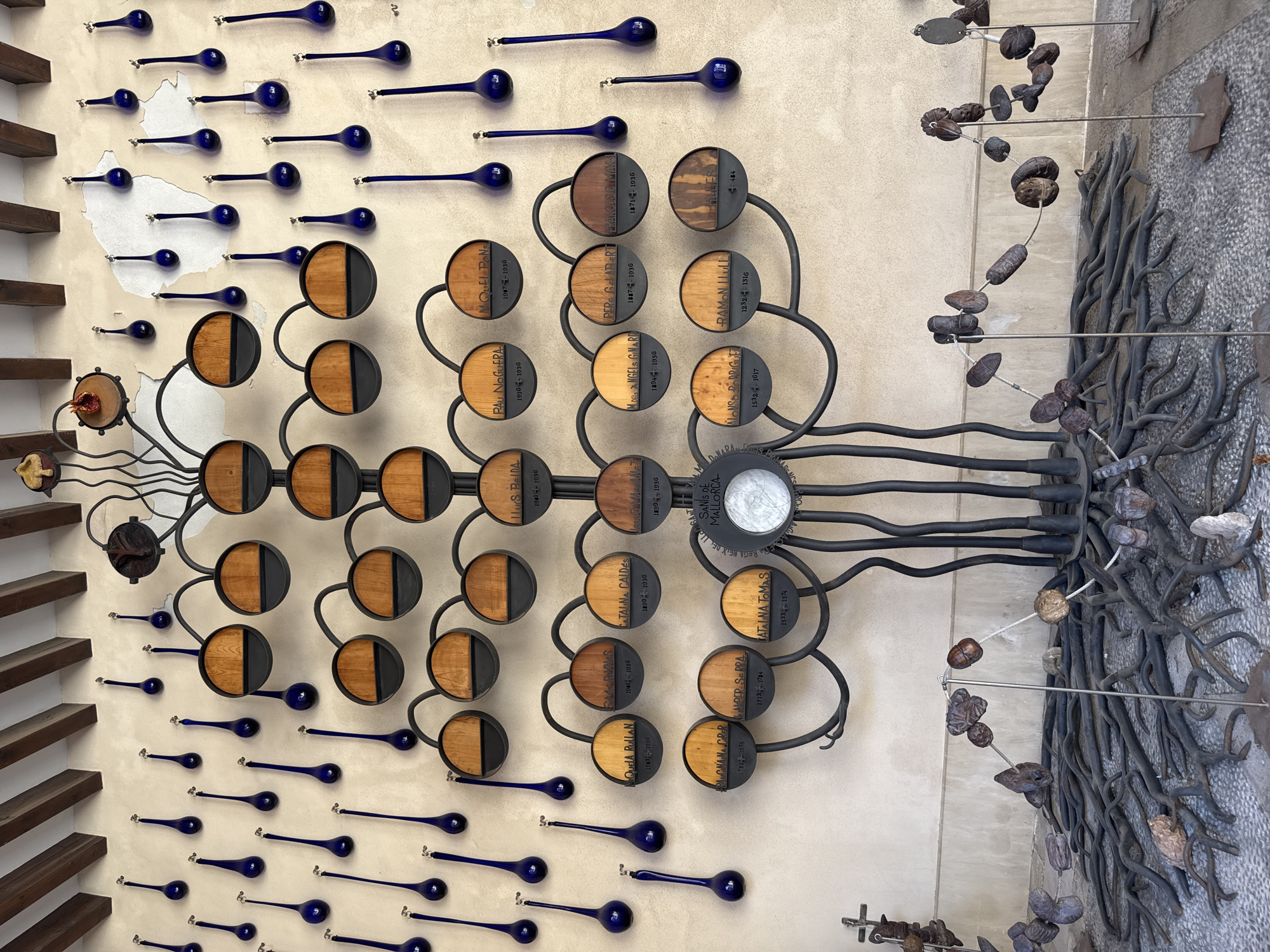 The image shows a sculptural installation resembling a tree, located outdoors in Palma de Mallorca, Spain. The "tree" is made of dark metal and features numerous circular wooden plaques mounted on its branches. Each plaque displays a name and a set of dates, suggesting a genealogical or historical significance. These plaques are arranged in rows and levels, mimicking the structure of a family tree.
Cascading down from the ceiling or upper wall, and also interspersed around the main tree structure, are numerous dark blue glass teardrop-shaped objects, each suspended by a thin wire and hook. These appear to be decorative elements.
At the base of the metal tree sculpture, the "roots" are also rendered in metal, fanning out across the ground. Scattered around these metal roots and on stands are various natural objects, such as stones and what appear to be dried seed pods or fruits, some strung together with wire, resembling a naturalistic artistic element.
The background is a textured wall with some peeling plaster and wooden beams visible at the top, suggesting an architectural setting. The ground is paved with small, multi-colored stones. The lighting suggests it is daytime, with diffused light and shadows indicating an overcast or partially shaded environment. There are no people visible in the image. The overall impression is an artistic representation of lineage and history, integrated into its surroundings.