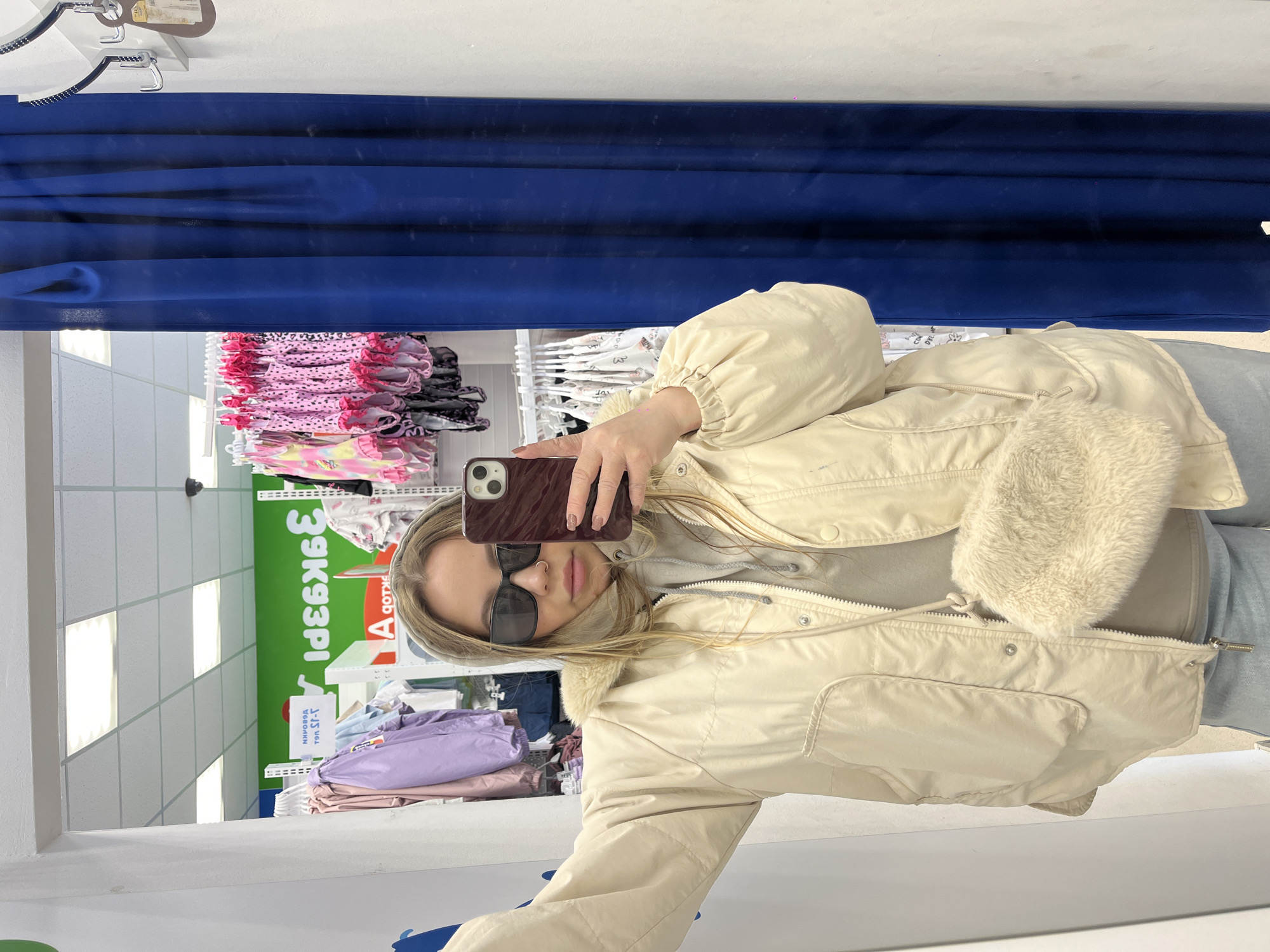 A young woman wearing sunglasses and a beige jacket with a fur collar is taking a selfie in a clothing store in Novopokrovskaya, Russia. The store appears to have a wide selection of clothing displayed on racks and shelves. The selfie is taken in front of a blue curtain.