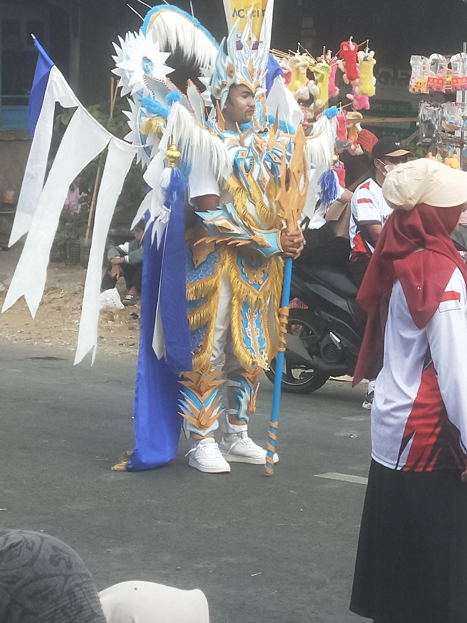 The image captures a street scene in Boja, Indonesia, likely during a parade or festival. The central figure is a person adorned in an elaborate, blue and gold costume, complete with a large headdress and feathered wings. They are holding a long, blue staff with gold accents and white ribbons are attached to their left side. The costume appears to be inspired by mythical or fantastical elements.
To the right of the central figure, another person is partially visible, wearing a red headscarf and a white shirt with red accents. They are looking away from the camera. In the background, several stalls with colorful items, possibly toys or souvenirs, are displayed. A motorcycle is parked behind the central figure. The ground is paved asphalt, indicating a street or road. The overall atmosphere suggests a lively public event.