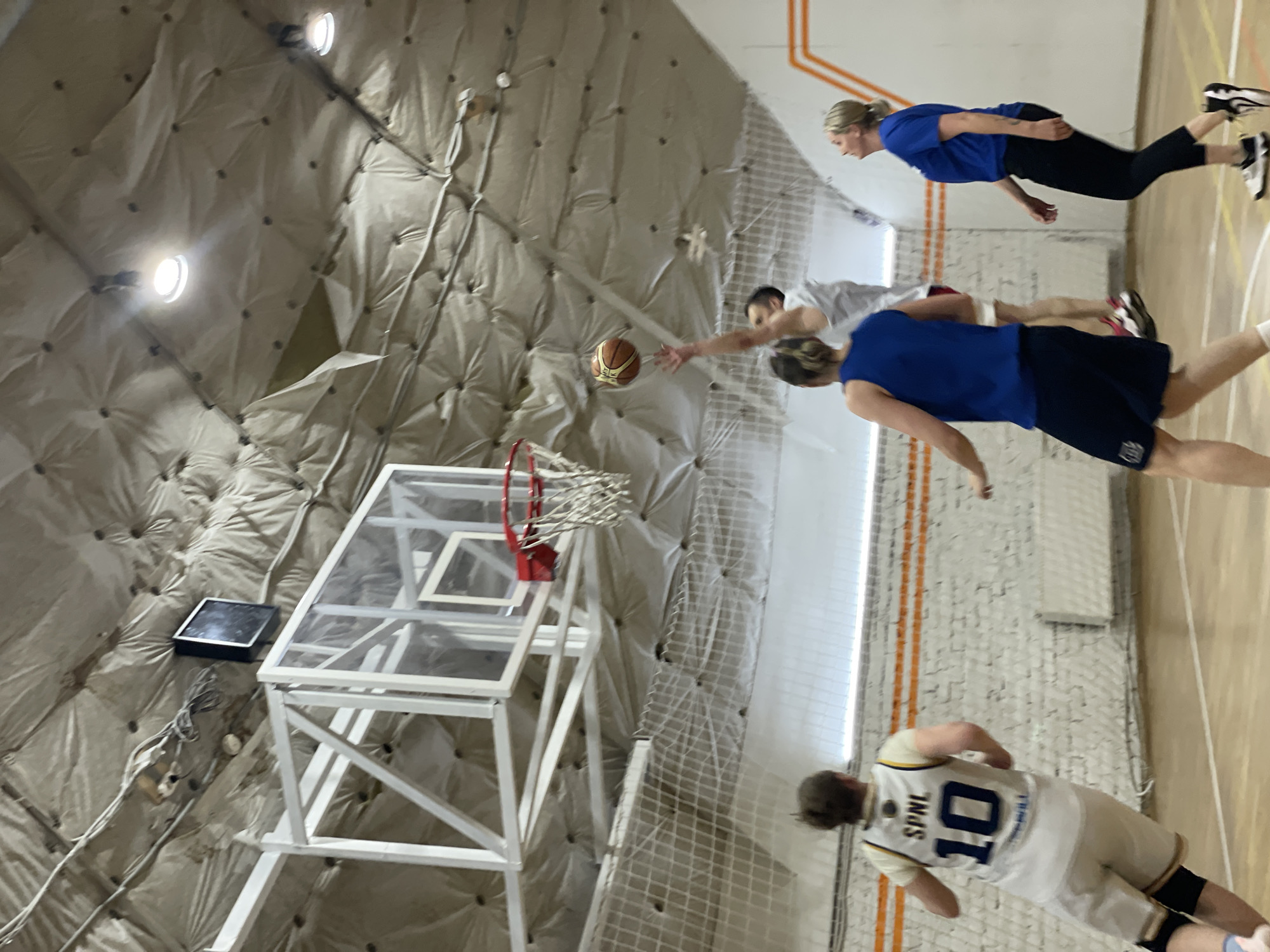 The image captures a basketball game in progress inside a gymnasium. A basketball is in the air near the hoop. Four individuals are actively participating in the game while another watches. One person in a white jersey marked with "SPNL 10" is also involved. The gym appears to be located in Saint Petersburg, Russia. The walls and ceiling of the gymnasium are covered in quilted padding. The floor is made of polished hardwood and features basketball court markings.