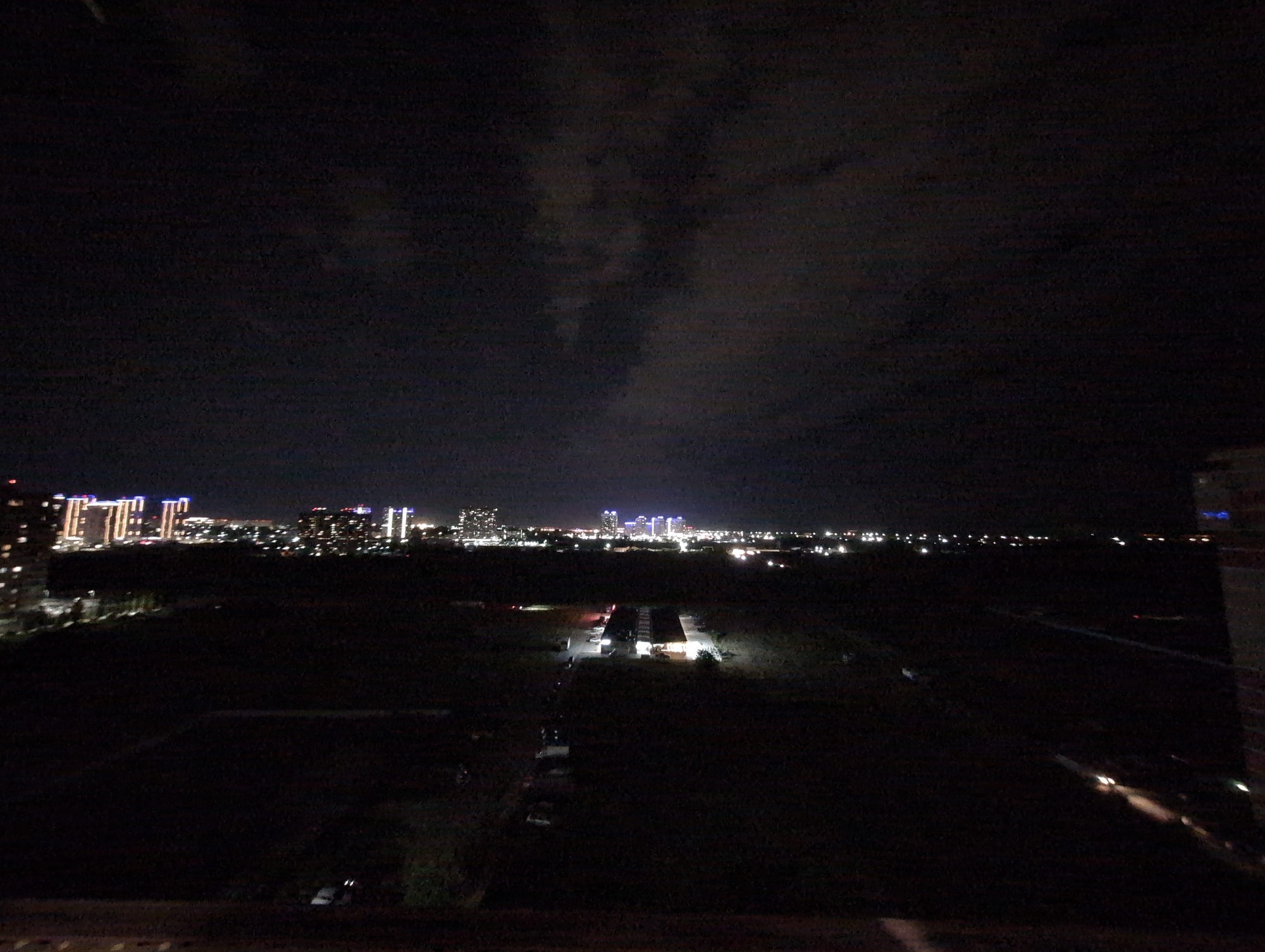 An aerial nighttime view of a city shows a skyline dominated by illuminated skyscrapers. In the foreground, a dark, flat area with some scattered lights suggests a park or undeveloped land. A few vehicles are visible, some parked and others with illuminated headlights, indicating movement on the roads. The overall impression is one of urban expansion and nighttime activity.
