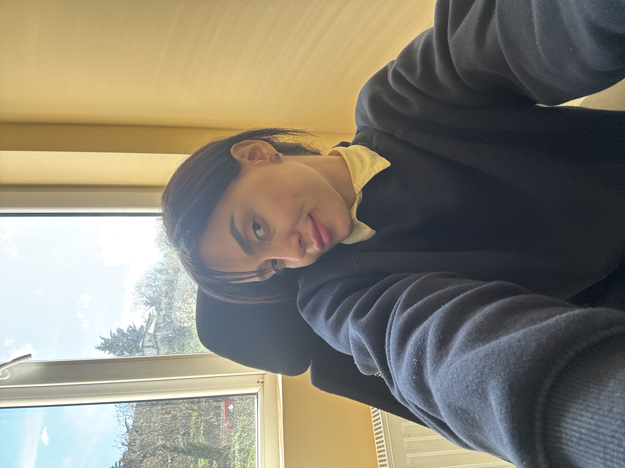A young person sits indoors by a window. The person is wearing a dark blue sweater over a yellow collared shirt. They are sitting in a black mesh office chair. The window looks out onto a partially visible outdoor scene. The scene appears to include trees and buildings under a cloudy sky. The location appears to be in Tbilisi, Georgia.