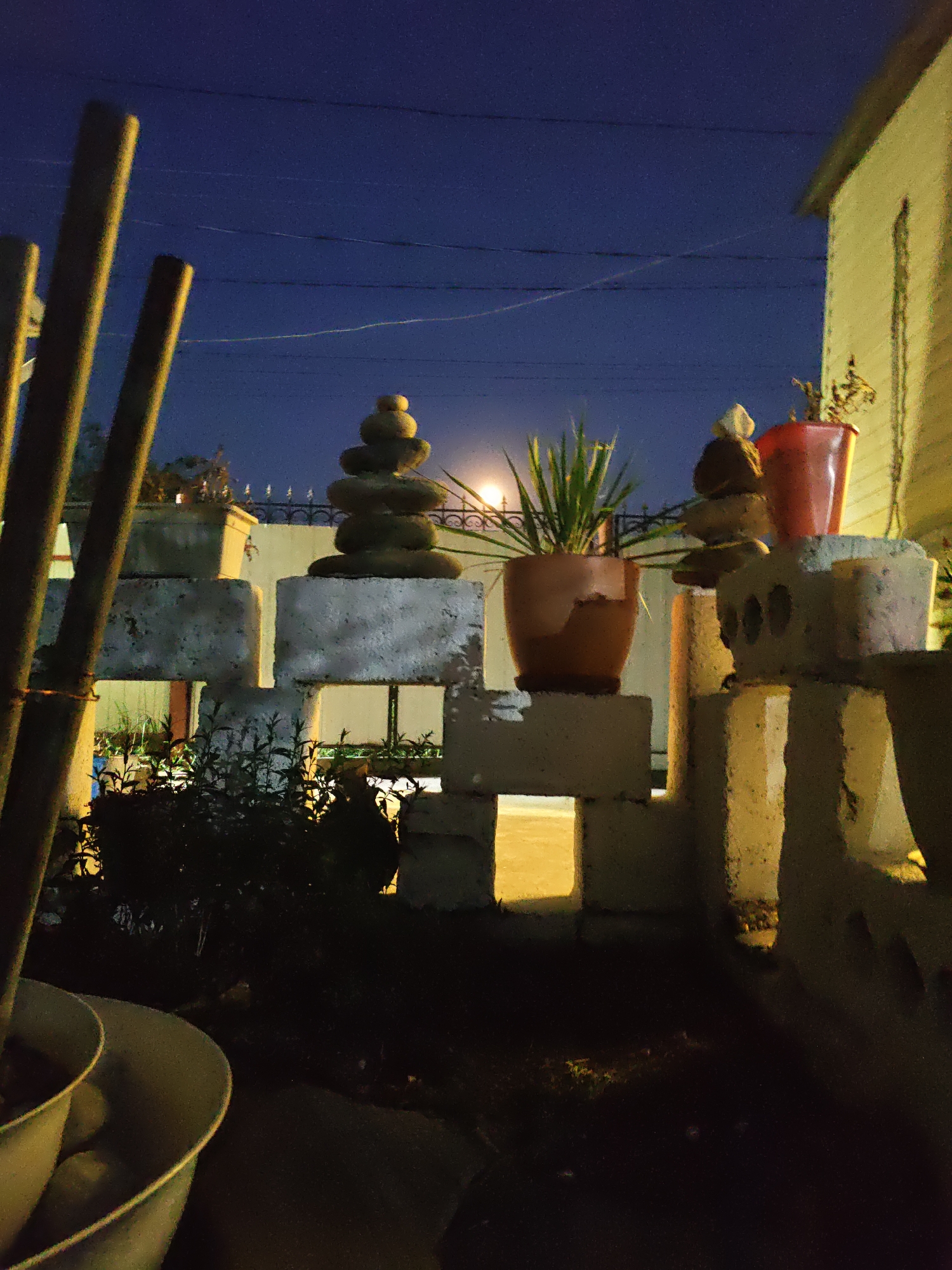 The image captures a nighttime scene in Rostov-on-Don, Russia. In the foreground, several large, weathered, hollow concrete blocks are stacked to form a low barrier or wall. Some of these blocks have openings that reveal a dimly lit space beyond, suggesting a yard or garden area. To the left, three vertical, dark-colored poles, possibly part of a fence or structure, are partially visible, out of focus. In the mid-ground, the concrete blocks continue to form a low wall. On top of these blocks, two potted plants are visible. One is a terracotta pot containing a spiky, green plant. Adjacent to it, a larger, red plastic bucket holds another plant, which appears withered. Stacked atop each other on another section of the concrete barrier are several smooth, rounded stones, resembling a cairn or Zen garden feature. The background shows a dark, clear night sky with power lines stretching across it. To the right, a portion of a building with light-colored siding and a sloped roof is visible, illuminated by an unseen light source. The overall impression is one of an outdoor space, possibly a backyard or patio, at night, with decorative elements and urban infrastructure present.