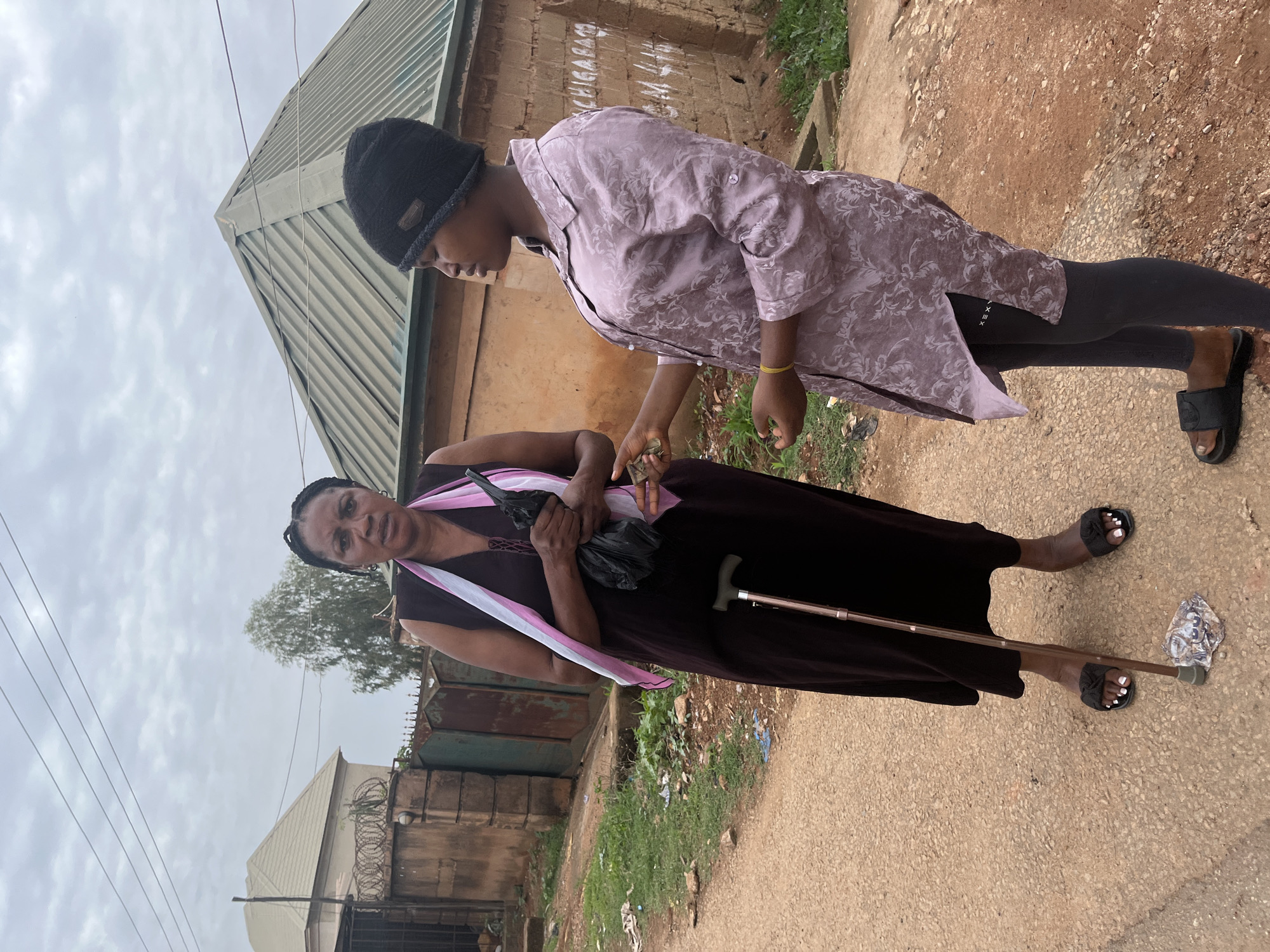 In Jos, Nigeria, a young woman in a lilac floral patterned top, gray leggings, and a black beanie stands facing a senior woman. The older woman is wearing a long black dress with a pink and white sash. She holds a black plastic bag and a silver cane. They stand on a dirt and paved road alongside buildings with metal roofs and brick walls.