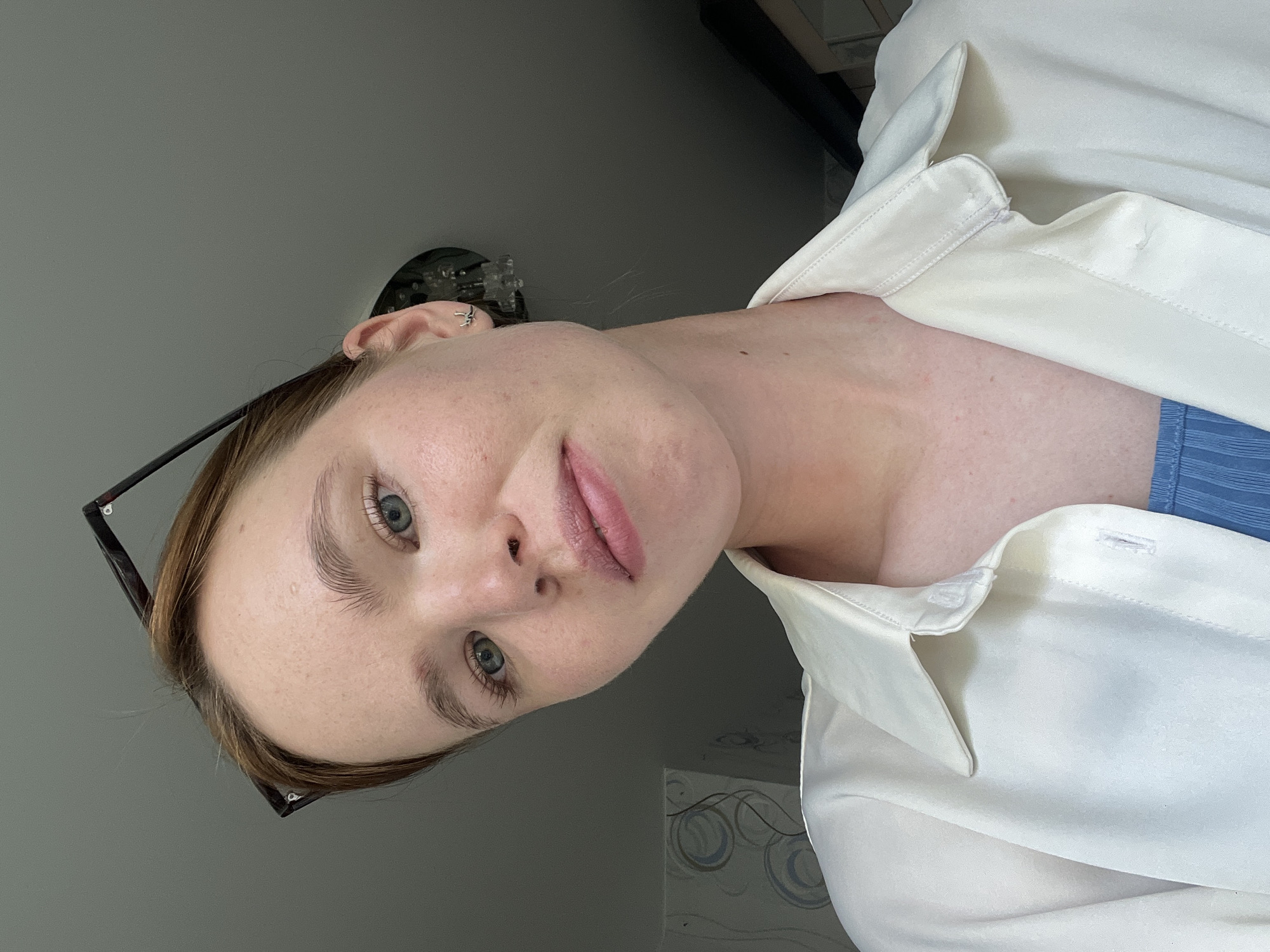 A young woman is shown in a close-up selfie. She has light skin, light-colored eyes, and light brown hair. She wears black-rimmed glasses. She is wearing a white, button-up shirt. A blue garment can be seen underneath. The background is a plain gray wall with a decorative object above her head and decorative swirls on the lower-right. The location is unknown, including whether it is in Chaikovsky, Russia.