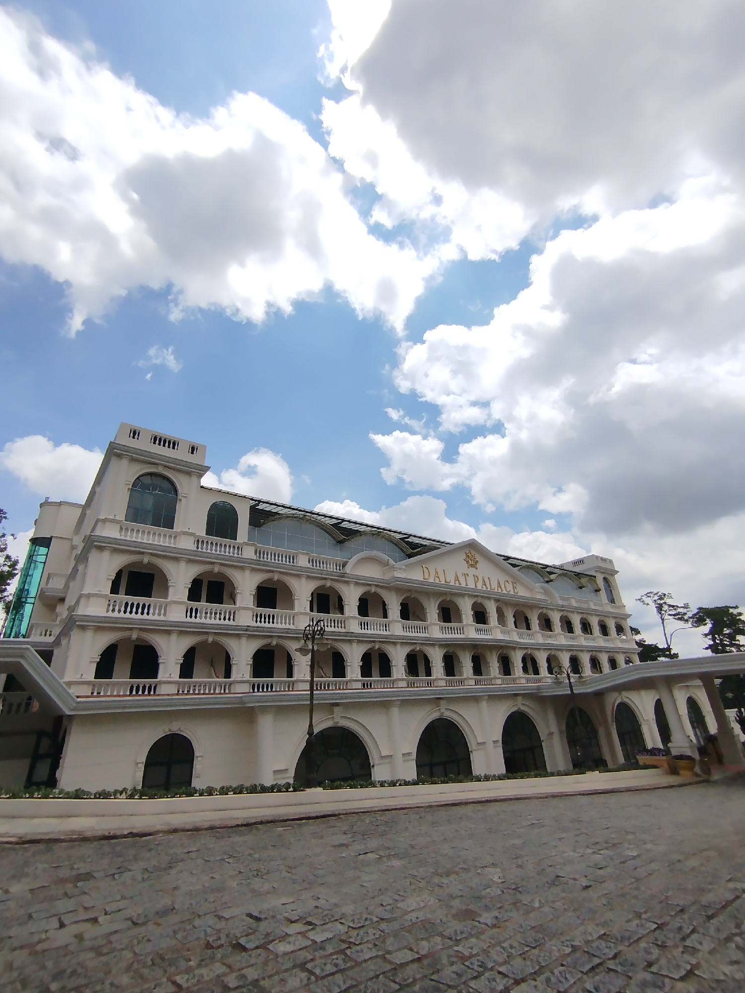 The image shows the Dalat Palace Hotel in Da Lat, Vietnam. The hotel is a white, multi-story building with arched windows and balconies. The name "DALAT PALACE" is inscribed in gold letters on the top of the building. A lamp post stands in front of the hotel. The sky is blue with white clouds. The foreground shows a cobblestone pavement.