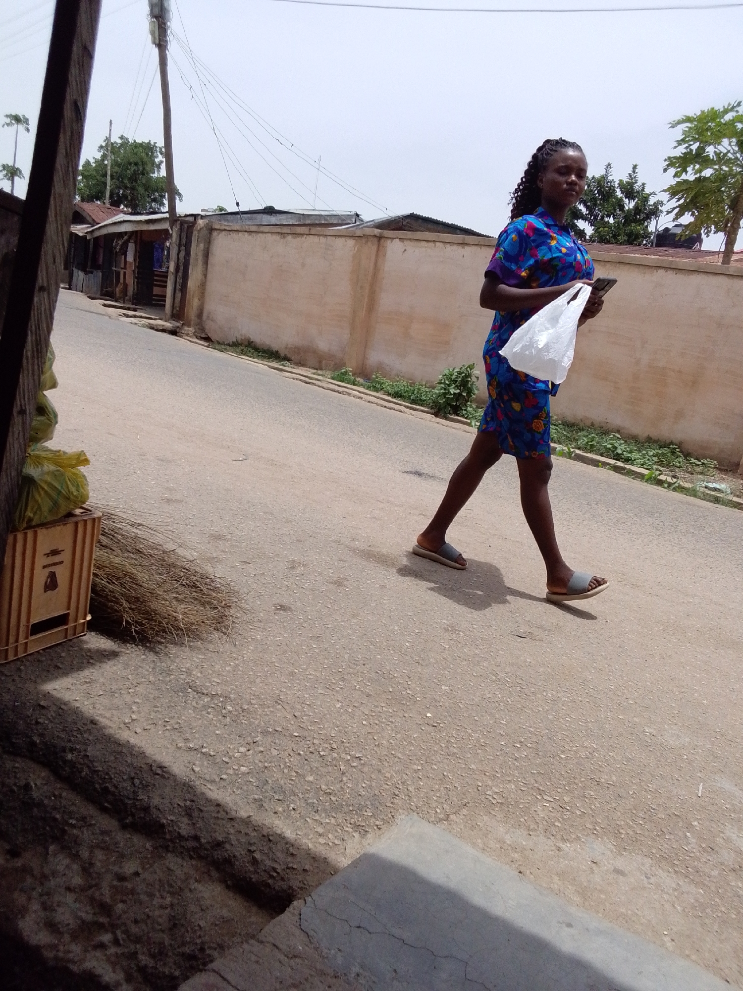 A young woman is walking on a paved road in what appears to be a residential or commercial area. She is wearing a patterned blue dress and sandals. She is carrying a white plastic bag and a mobile phone. The road is bordered by buildings and a wall, with some vegetation growing along the wall's base. In the foreground, a wooden crate with what looks like trash bags on top, sits next to a bundle of brooms, which are partly in shadow. Utility poles are also visible. The scene appears to be in the city of Jos, Nigeria.