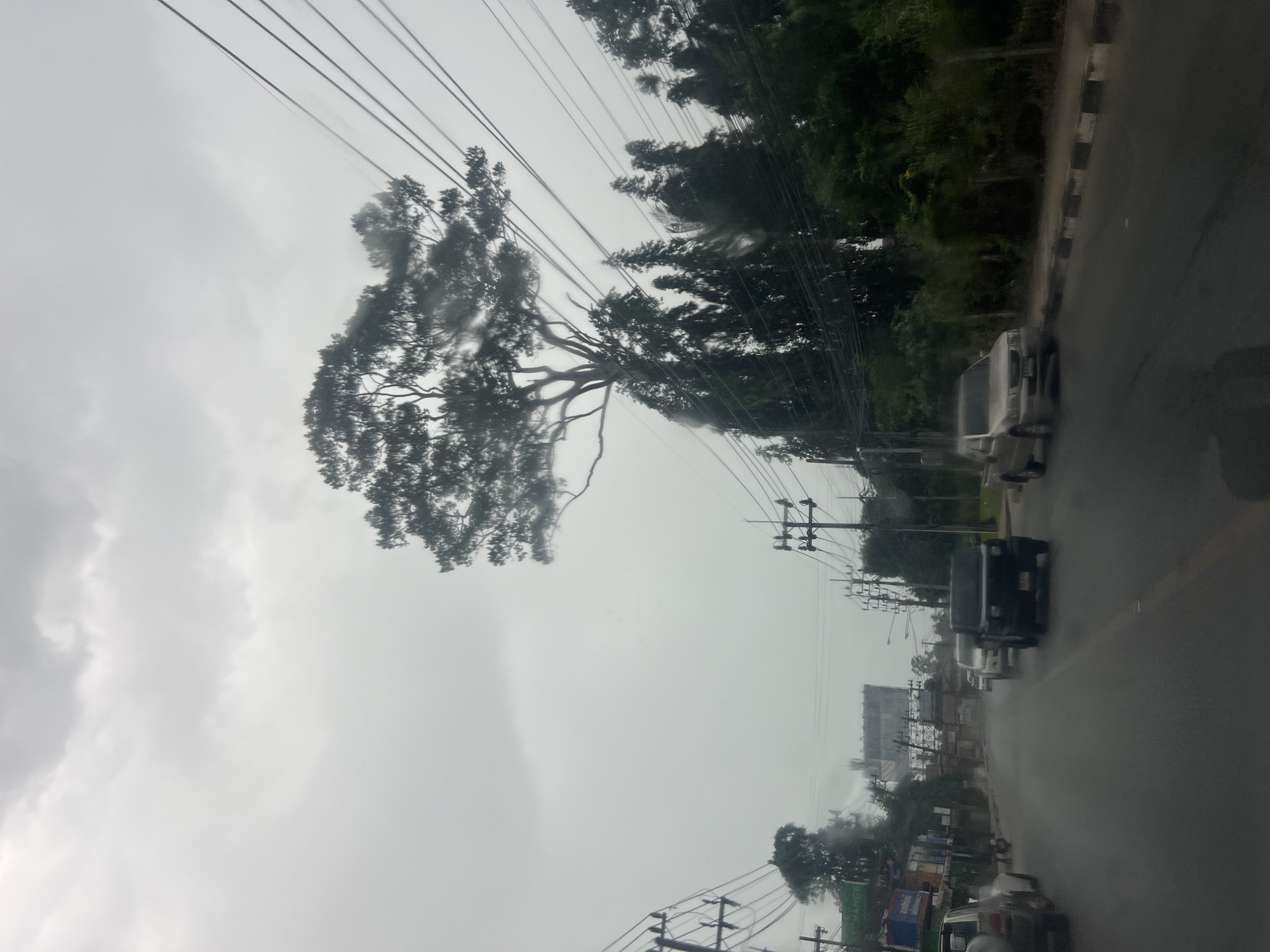 The image depicts a street scene in the city of Ko Samui District, Thailand, during a rainstorm. The sky is overcast and gray, with heavy clouds. Raindrops are visible on the camera lens, creating streaks across the image. The road is wet and reflective, with several vehicles driving in the rain. In the foreground, a silver pickup truck is parked on the right side of the road. Further down the road, a black SUV and other cars are moving in traffic. On the left side of the road, buildings and shops line the street, with power lines and utility poles overhead. A large tree with sparse foliage stands prominently on the right side of the road, its branches extending towards the sky. The overall atmosphere is somber and wet, characteristic of a tropical downpour.