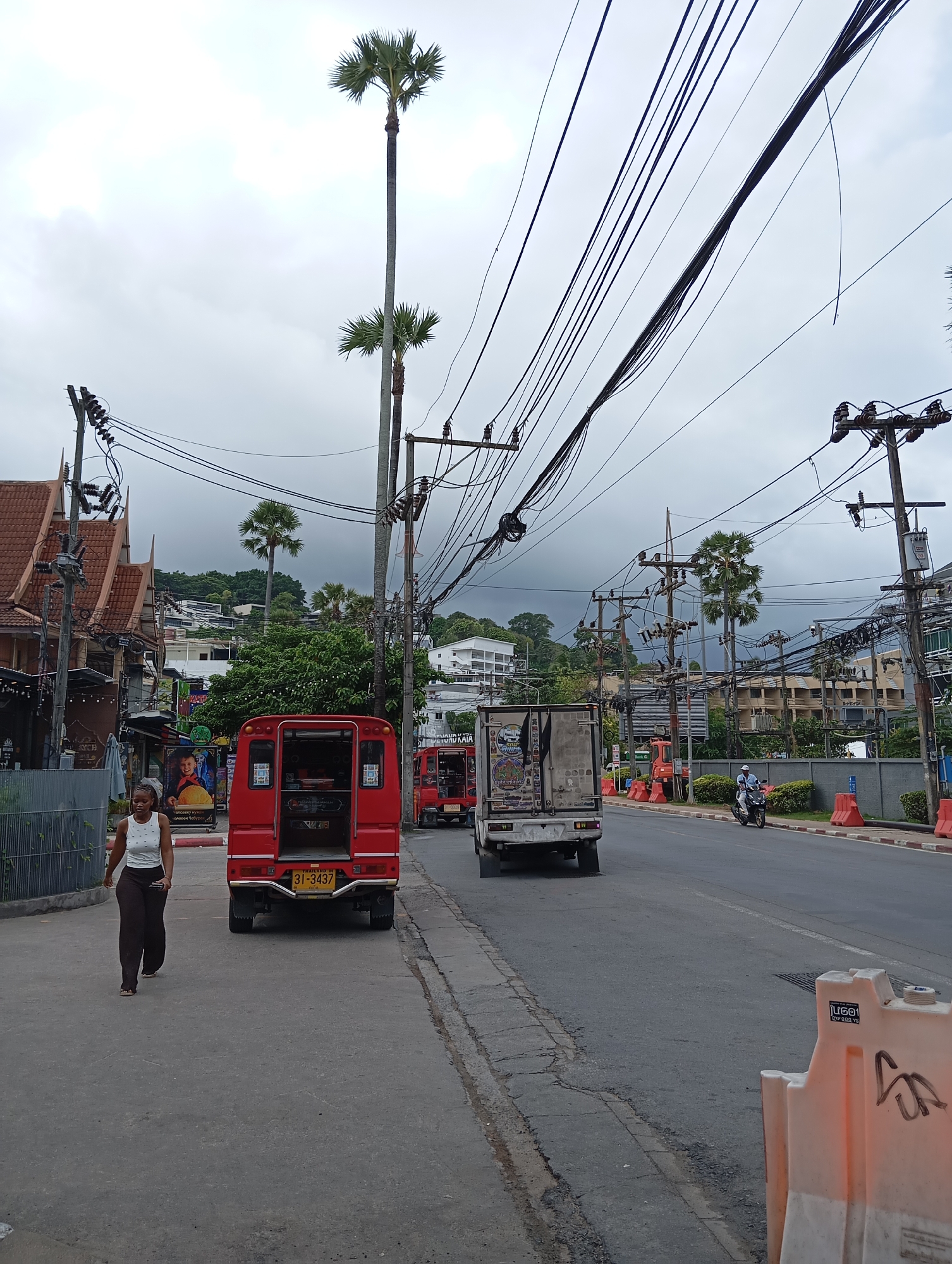 The image shows a street scene in Karon, Thailand, under an overcast sky. A young Black woman walks on the sidewalk on the left side of the road. She is wearing a white tank top and dark pants. To her right, a red open-back tuk-tuk is parked, with its license plate "31-3437" visible. Further down the street, other vehicles, including a white truck and another red tuk-tuk, are present. The road is lined with buildings and palm trees. Overhead, a dense network of electrical wires is strung between poles, creating a cluttered visual effect against the sky. Orange traffic barriers are placed along the right side of the road.