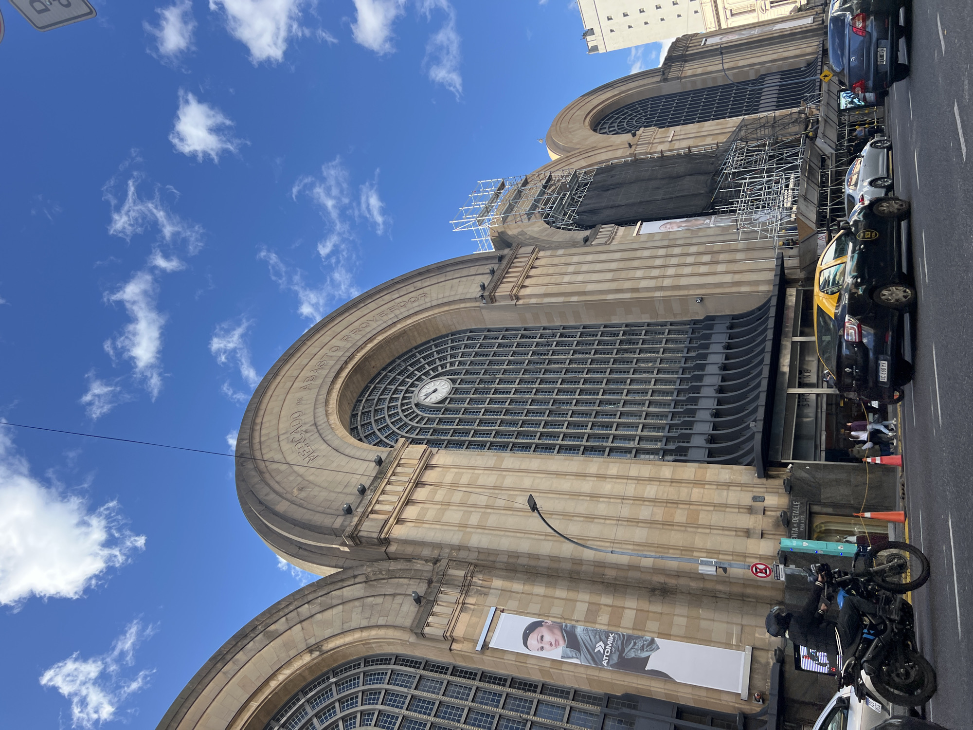 The image captures a view of the Mercado de Abasto, located in Buenos Aires, Argentina. The architecture is visible, including the arched windows and stonework, with a large clock visible in the upper arch. Scaffolding is present on the building's facade, indicating ongoing maintenance or restoration. Street-level elements include cars, a motorcycle with a person on it, and pedestrian activity near the entrance of a shopping area. Signs and a poster for "Atomik" are also visible on the building's exterior. The sky is blue with scattered clouds.