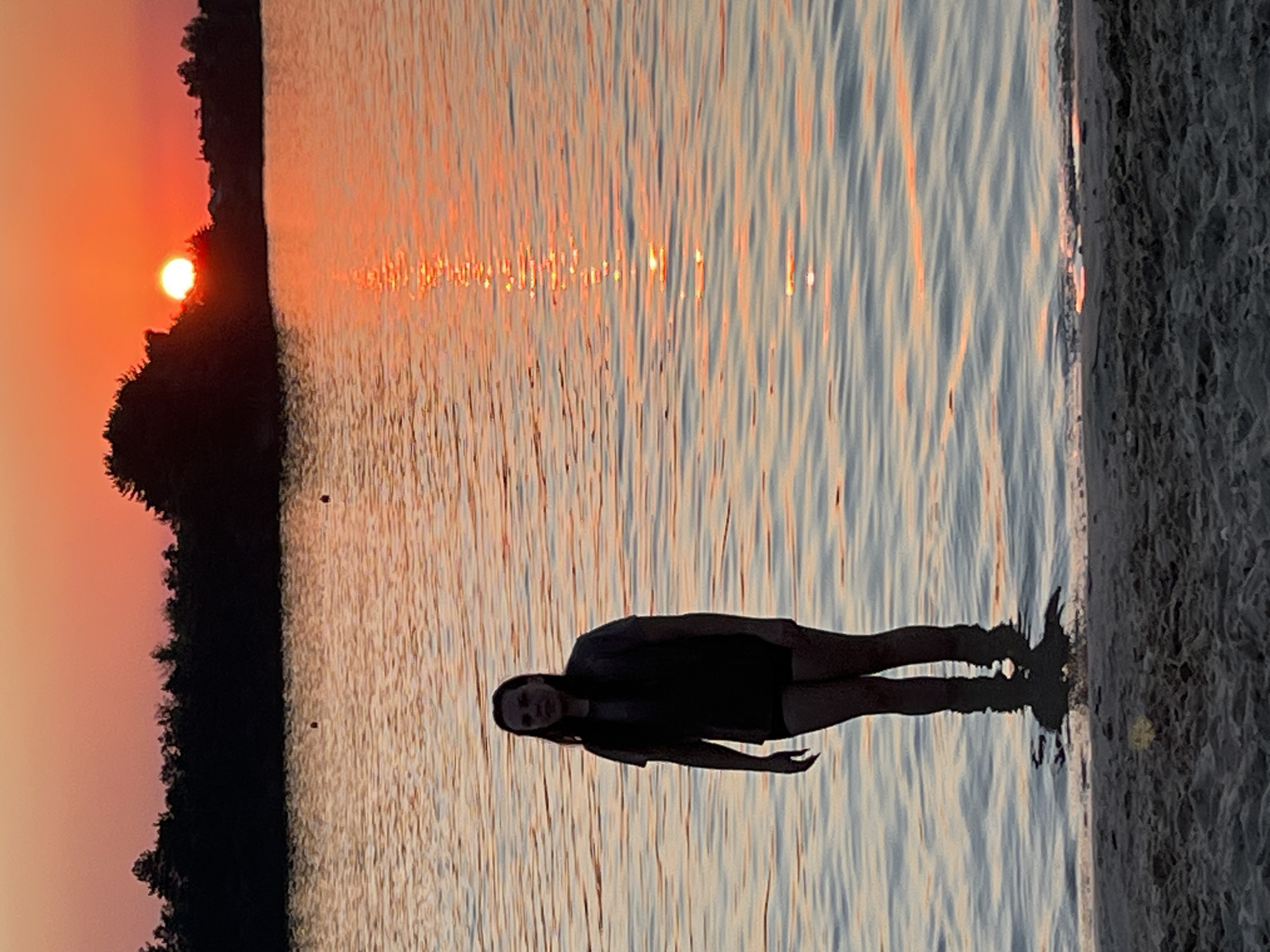 A full-body shot shows a young woman standing in shallow water at the edge of a sandy beach, with a sunset visible in the background. The woman, who has dark hair and is wearing a dark t-shirt and shorts, is looking towards the camera. Her legs are submerged up to her knees in the water, and her shadow is cast on the sand beneath the water. The water itself reflects the warm orange and red hues of the sunset, creating a shimmering effect on the surface. In the distance, a line of dark trees forms the horizon, silhouetted against the vibrant sky. The sun is a bright, glowing orb, partially obscured by the trees on the right side of the frame. The scene appears to be a tranquil evening at a beach or lake. The location is identified as Bezliudivka, Ukraine.