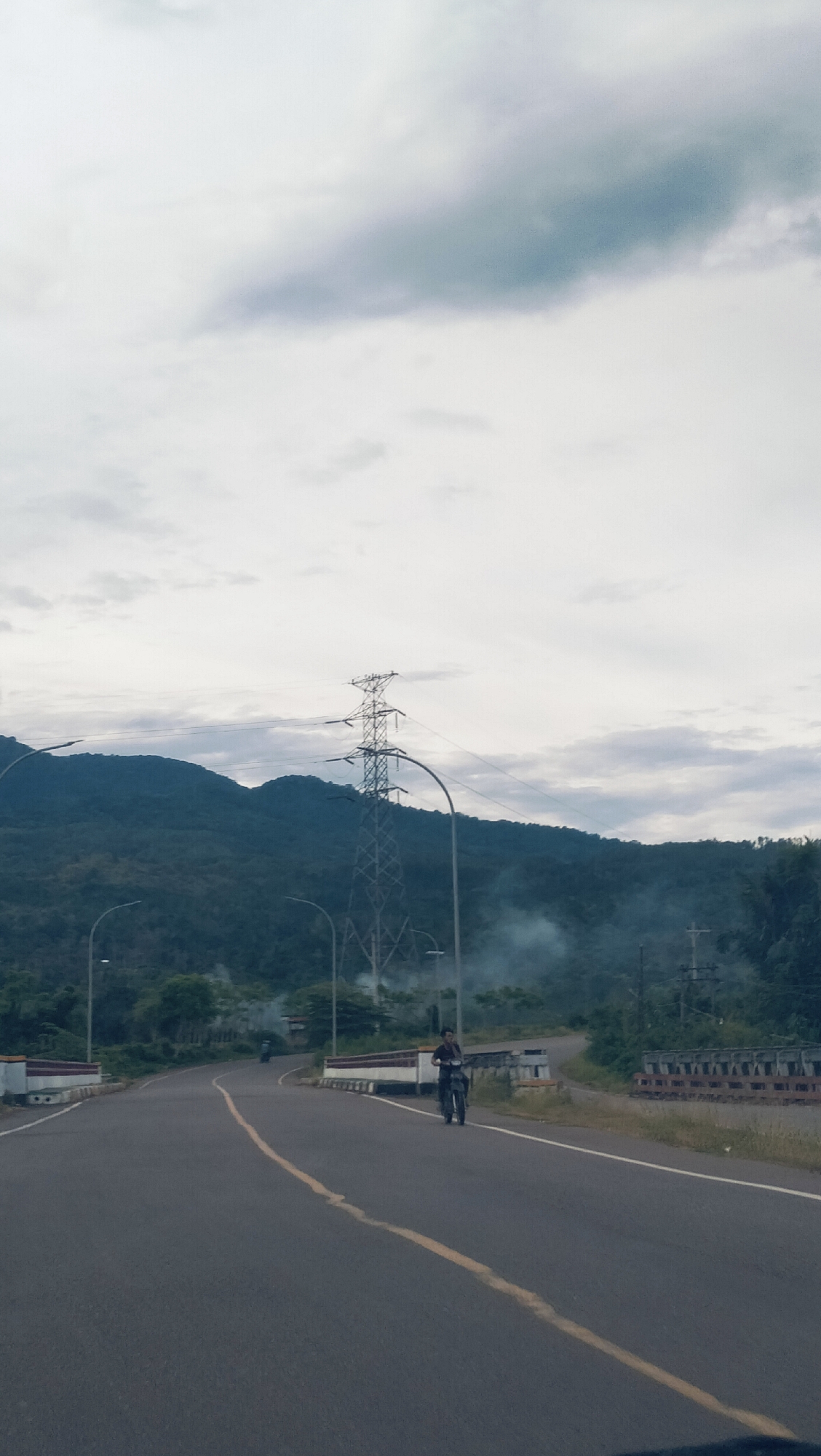 This vertical full shot depicts a rural road cutting through a mountainous landscape in Lamtamot, Indonesia, under an overcast sky. The asphalt road dominates the foreground, featuring a distinct double solid yellow line running down its center. A person, dressed in dark clothing, walks on the right shoulder of the road, appearing to carry an item across their chest. Further along the road on the left, a small motorbike is faintly visible in the distance.
The road is bordered by low concrete and metal railings, suggestive of a bridge or elevated section, and modern street lights with curved poles illuminate the path. In the mid-ground, a tall metal electrical transmission tower stands prominently against the backdrop of dense, green forested mountains that stretch across the horizon. Wisps of white smoke rise from the right side of the image, originating from behind the road, possibly indicating a small fire or controlled burning.
The sky is heavily clouded with varying shades of grey and light blue, creating a diffused light that suggests an overcast day. The scene conveys a sense of a quiet, remote environment where nature and essential infrastructure coexist. There is no visible text in the image.
