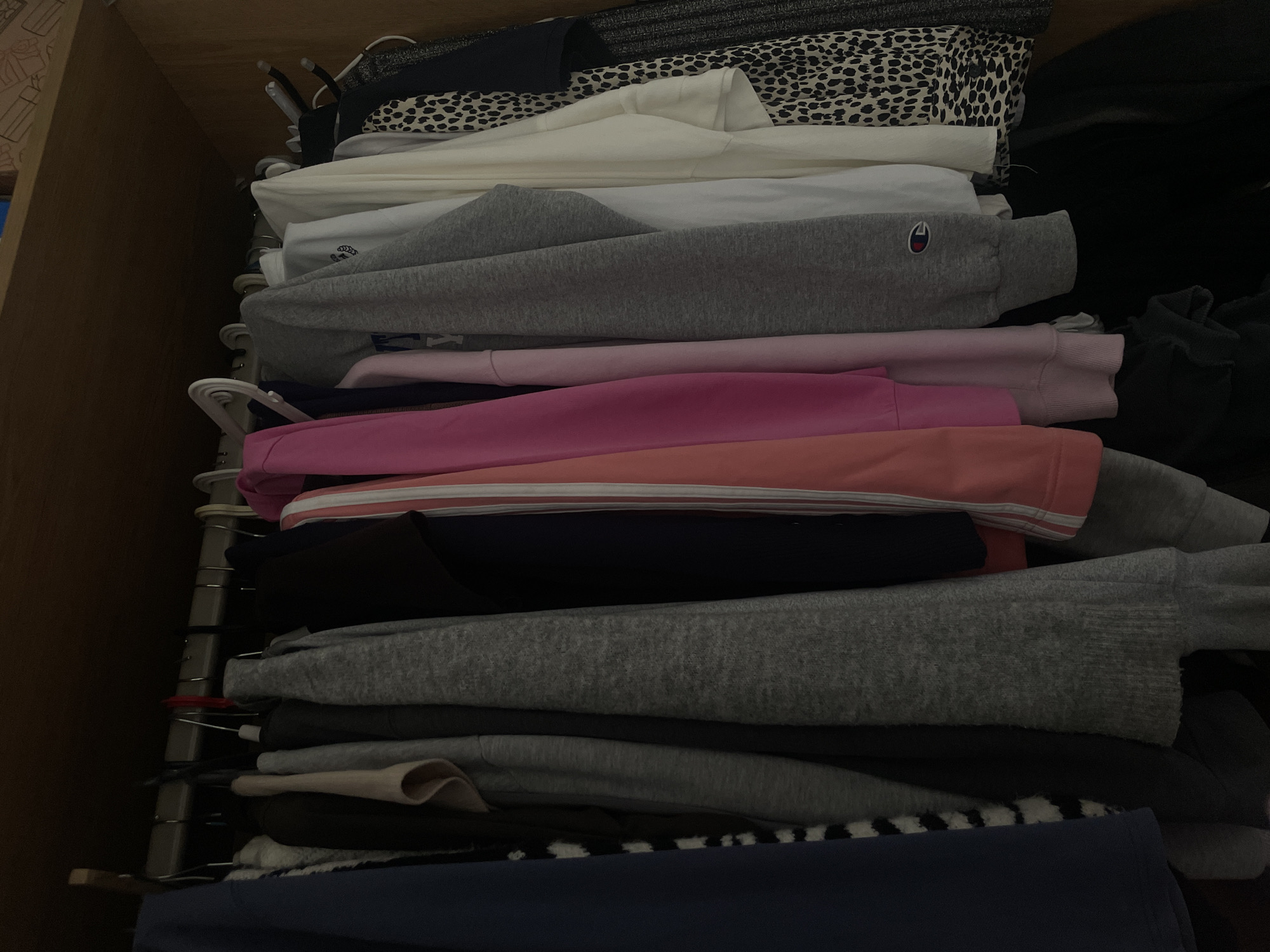 The image displays a vertical stack of neatly folded clothes inside what appears to be a wooden wardrobe or deep drawer. The garments are primarily sweaters, hoodies, and long-sleeved shirts, arranged in horizontal layers.
The color palette of the clothing includes various shades of grey, light and dark pink, white, dark blue, and black. Near the top, there is a piece of fabric with a black and white animal print pattern, resembling leopard or dalmatian spots. On the left side of the stack, several white plastic hangers are visible, attached to a vertical metal rod or shelving system, suggesting an organized storage space.
A notable detail is the distinct red and blue Champion "C" logo embroidered on a grey hoodie or sweatshirt, positioned in the upper-middle section of the stack. Another small, light-colored embroidered design, possibly "G+R" or similar, can be faintly seen on a white garment directly above the Champion item. The overall lighting is dim, indicating an indoor setting. There are no people present, and the scene depicts organized clothing storage.
