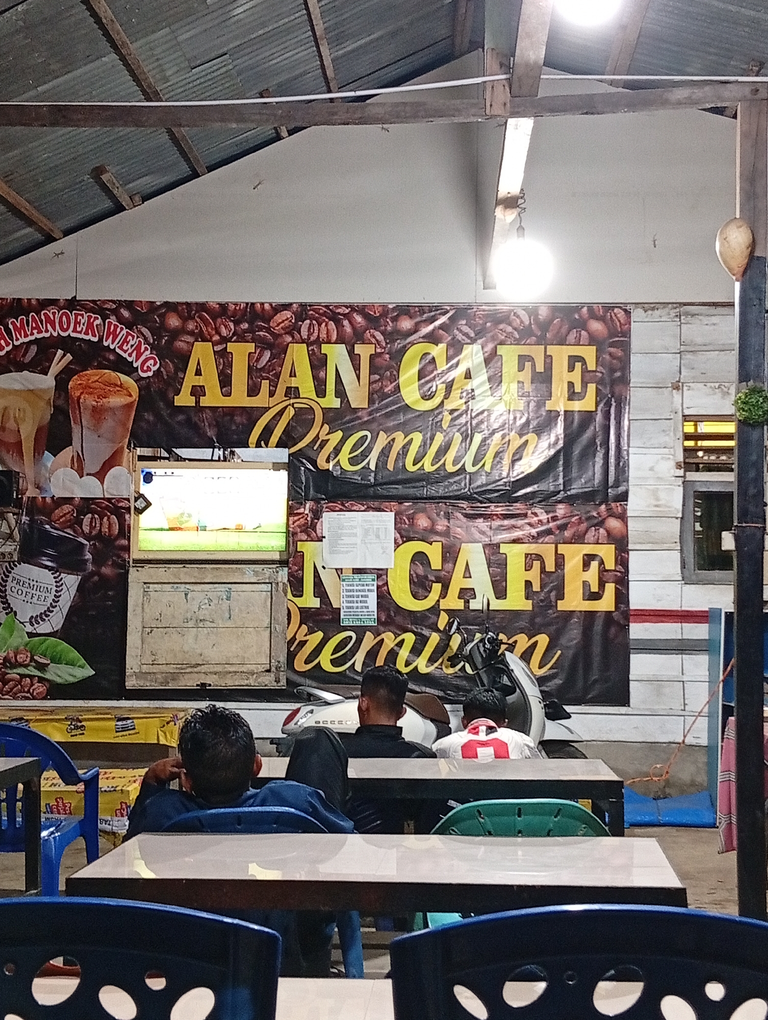 The image depicts an indoor cafe identified as "ALAN CAFE PREMIUM MANOEK WENG" in Lhokseumawe, Indonesia. The scene is illuminated by multiple bare incandescent light bulbs and a fluorescent tube, indicating low ambient light conditions.
The upper portion of the image reveals a corrugated metal roof supported by exposed wooden beams. A white-painted wall section is visible below the roofline, with three small lizards observed on its surface. A large, dark banner advertising the cafe dominates the background, featuring coffee beans as a motif and yellow text reading "ALAN CAFE PREMIUM" twice, along with "MANOEK WENG" partially visible on the left. The banner also displays illustrations of coffee beverages and a "PREMIUM COFFEE" logo.
Centered on the banner, a bright rectangular screen, possibly a television or monitor, displays a green field, suggesting a sports event or game. Below the screen is a wooden structure, appearing to be a service counter or enclosure. To the right of the banner, a wall constructed of horizontal white planks is visible, featuring a window with yellow-tinted light within. A dark vertical wooden post is present on the far right, supporting a small green spherical decorative plant and a hanging, light-brown, gourd-like object.
In the mid-ground, three individuals are seated at tables with dark frames and reflective light-colored surfaces. The person closest to the camera is seated with their back to the viewer, wearing a dark blue shirt and dark pants, with their right hand near their head. Behind and slightly to their right, another individual wears a white and red jersey with the number "11" on the back. A third individual's head, wearing a dark shirt, is partially visible to the left of the person in the jersey. A white motor scooter is parked behind the seated individuals. Blue and green plastic chairs are arranged around the tables. Stacked yellow containers are visible behind a blue chair on the left. Electrical wiring runs along the wooden ceiling structure.