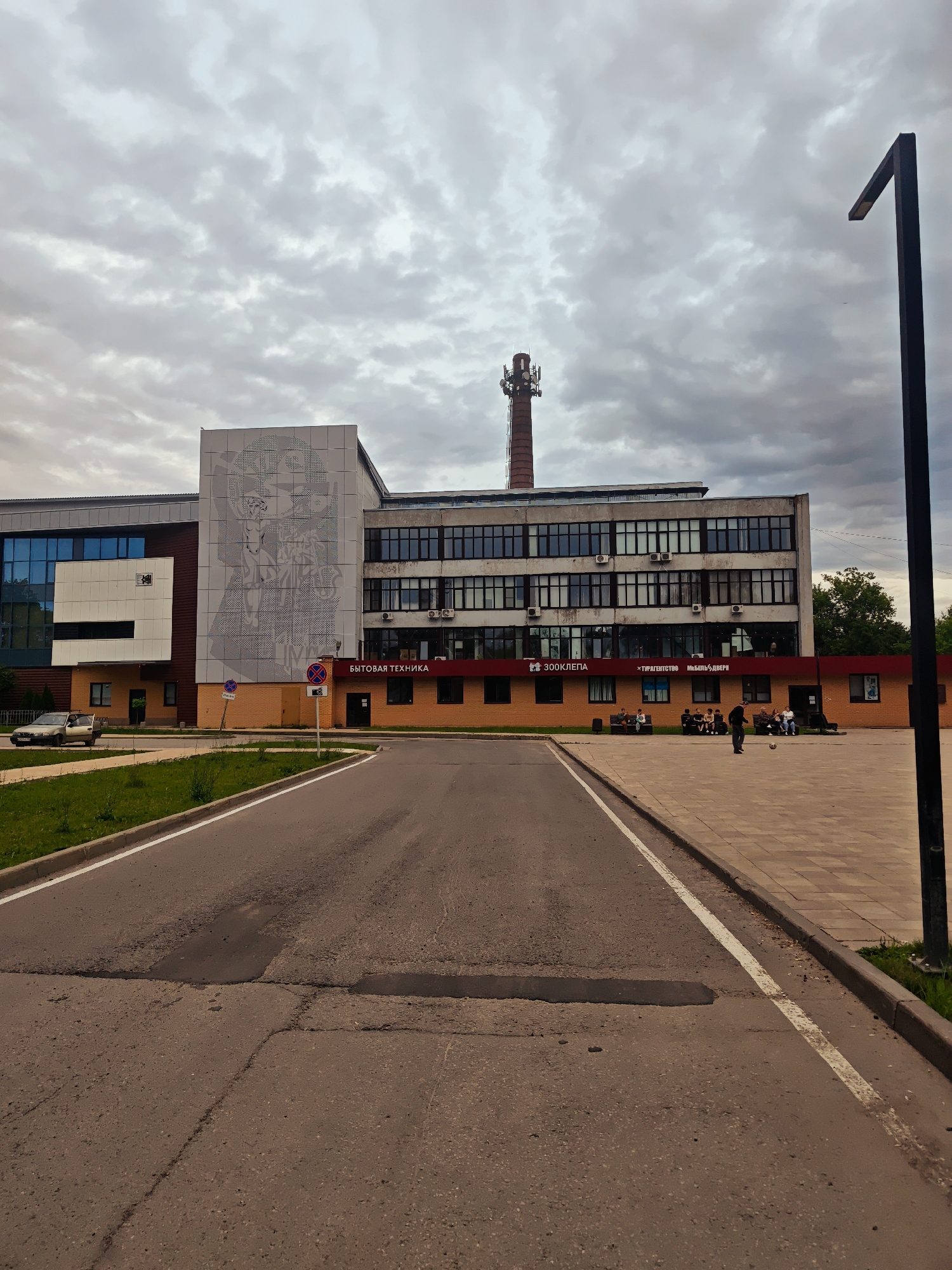 This media file depicts an overcast day in Zaprudnya, Russia, with a large, multi-story building dominating the scene. The building's facade features a mix of modern glass sections and older brickwork. A prominent feature is a large, grey, geometric mural on the upper part of the building, left of center. To the right of the mural, the building has multiple floors with rows of windows and visible air conditioning units. A tall, red brick chimney with telecommunication antennas on top stands behind the building.
In front of the building, a paved area functions as a plaza. Several people are gathered here, some sitting on benches, while a few individuals are standing. One person is in the process of kicking a soccer ball in the plaza, suggesting a casual or recreational activity. There is a paved road leading towards the building, with a grassy verge along its left side. The road appears somewhat worn, with visible patches and cracks.
Visible text includes "БЫТОВАЯ ТЕХНИКА" (Household Appliances) and "ЗООКЛЕПА" (Zoo Klepa), indicating the nature of businesses or services housed within the building. Further text reads "ТУРАГЕНТСТВО МЕБЕЛЬ ДВЕРЬ" (Travel Agency Furniture Door). Two traffic signs, one a "No Parking" sign and another with a circle and a diagonal line, are also present. The overall atmosphere is subdued due to the cloudy sky, suggesting it is likely daytime but not particularly sunny.