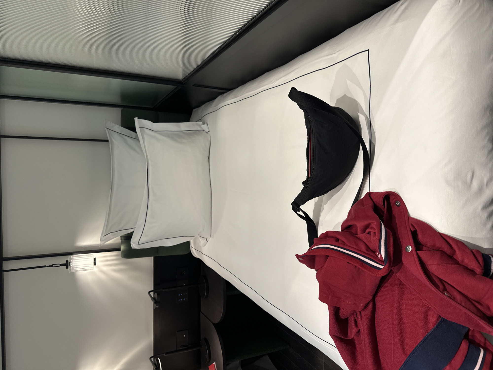 A bedroom in Tbilisi, Georgia, contains a bed with white linens, two white pillows, a black bag, and a red jacket with white and navy blue stripes.