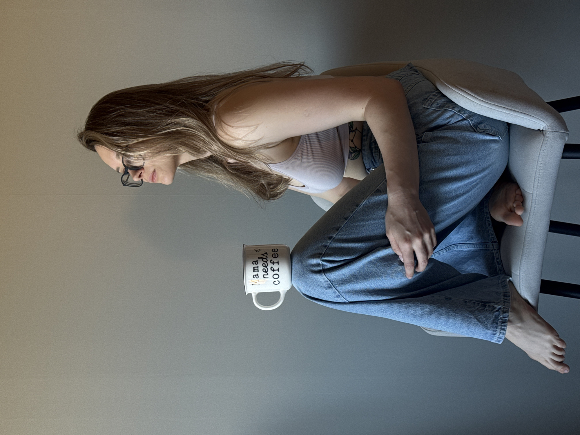 A woman is sitting in a chair, with a coffee mug on her lap. The mug reads "mama needs coffee". She is wearing glasses, a light-colored top, and blue jeans. She is barefoot. The chair has a white cushion and black legs. The background is a light gray wall. The location is not discernible from the image, therefore it cannot be determined to be Chaikovsky, Russia.