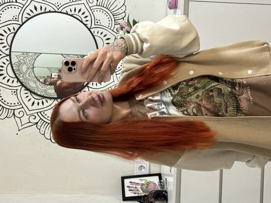 A young woman with long, vibrant red hair takes a selfie in a mirror. She wears a tan jacket with white trim over a printed t-shirt. The phone case is pink. Behind her is a white wall with a large, ornate mandala design and a round mirror reflecting a duplicate image of her. Below, a framed print and other decorative objects are visible. The overall impression is casual and artistic.
