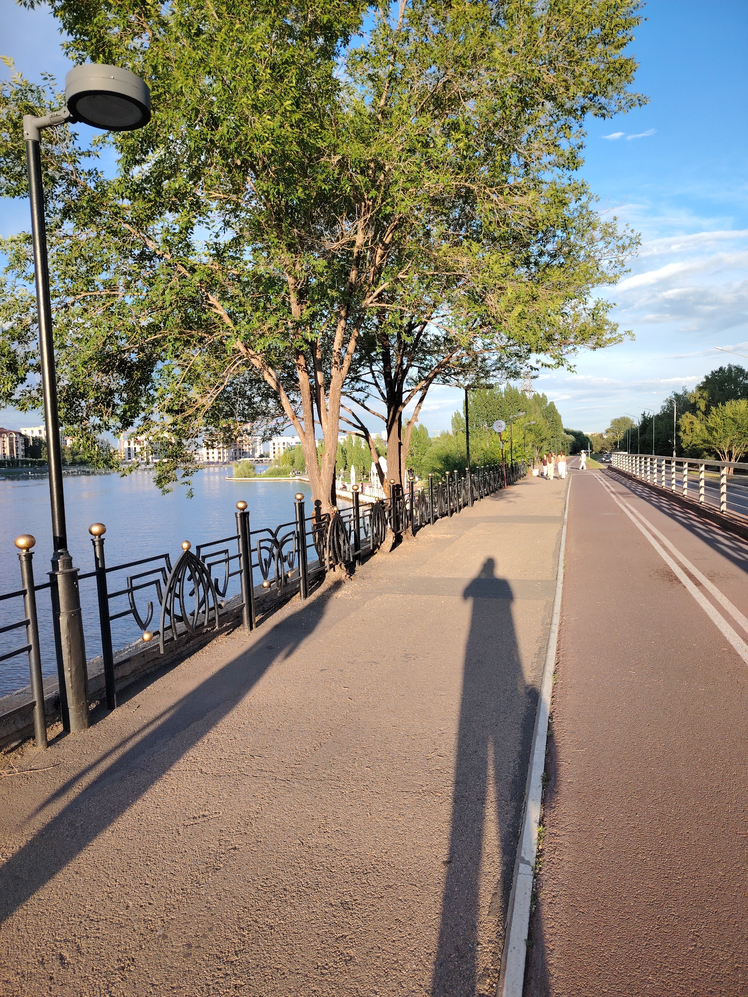 This image depicts a scenic view of the embankment along the Ishim River in Astana, Kazakhstan. The foreground features a paved walkway and a dedicated bicycle path, separated by a concrete curb. A prominent shadow of a person, presumably the photographer, is cast across the walkway, indicating a sunny day. To the left, a black lamppost with a modern design stands tall, and an ornamental black metal fence with golden spherical accents lines the edge of the river. Beyond the fence, the calm blue water of the Ishim River is visible, with buildings and trees lining the opposite bank. Several people are seen walking along the embankment in the middle ground, and a white car is parked further down the road on the right. The background reveals lush green trees and a clear blue sky with scattered white clouds, suggesting a pleasant day for outdoor activities.
