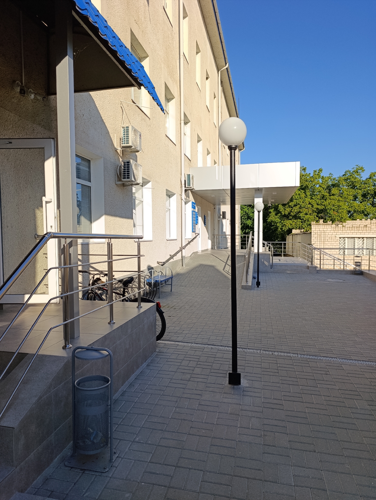 The image shows the exterior of the Dubăsari town council building in Moldova. A paved walkway leads up to the building's entrance, which is accessible by a ramp and stairs. The building has a light-colored facade and is equipped with air conditioning units. A blue awning covers the entrance. A black lamppost with a spherical white light fixture stands near the entrance. In the foreground, there is a metal trash can. Several bicycles are parked near the stairs. The area is well-lit by the sun, indicating it is daytime. Trees and a brick building are visible in the background.