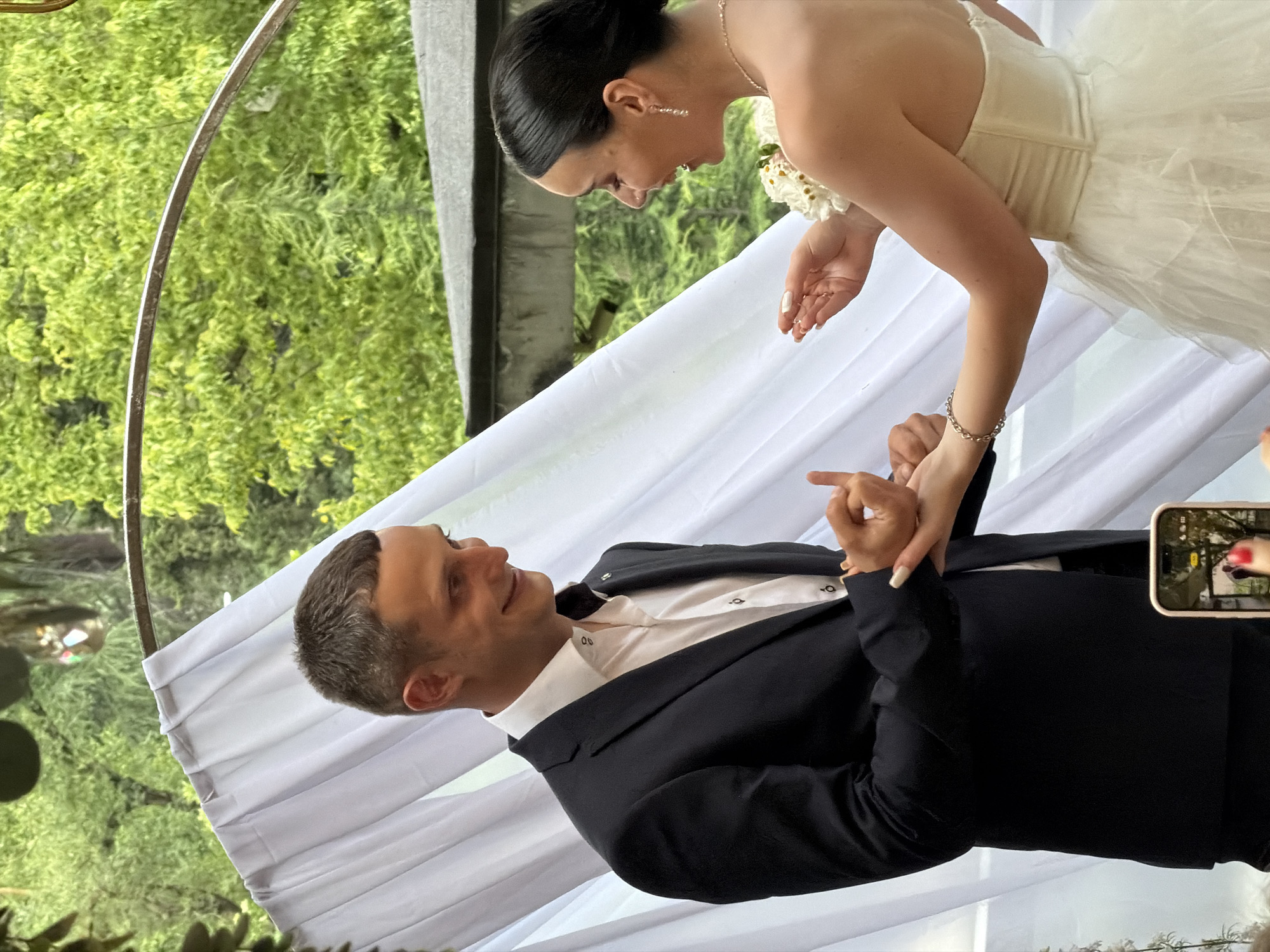 In Tbilisi, Georgia, at an outdoor wedding, a man in a dark suit and a woman in a white wedding dress stand facing each other. The man, with short brown hair, wears a white shirt and a dark suit jacket, smiling as he holds the woman's left hand and makes a gesture with his right hand. The woman, with dark hair pulled back, wears a strapless wedding dress and smiles as she holds out her hands, one of which is touching the man's. She wears earrings and a bracelet. A white drape forms a backdrop behind them, with lush green trees and foliage visible in the background. A metallic archway is also present. In the foreground, a smartphone is being used to record the scene.