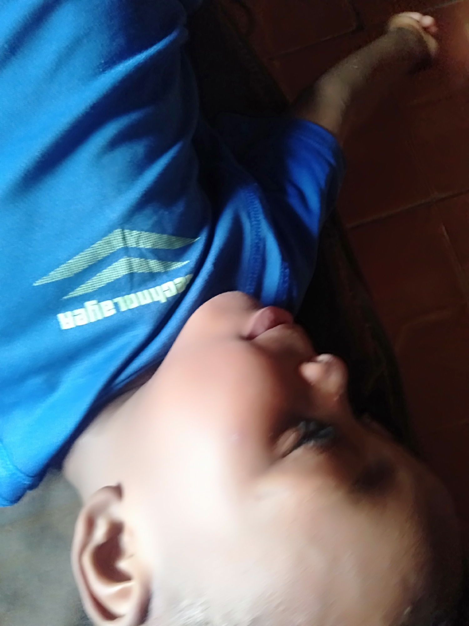 A young person, likely a child with dark skin, is depicted lying on their side on a reddish-brown tiled floor. The child is wearing a bright blue short-sleeved t-shirt. Their head is tilted back slightly, with an ear, cheek, and lips visible. Their eyes appear closed or mostly closed, suggesting they are sleeping or resting peacefully. An arm is extended towards the right side of the frame, with a hand that seems to be loosely clenched.
The blue t-shirt features light green chevron-like patterns and inverted white text that, when upright, reads "technolayer". The floor is composed of square tiles with visible grout lines. The lighting appears somewhat dim, indicating an indoor setting. The overall impression is of a child at rest in a casual home environment in Shangisha, Nigeria.