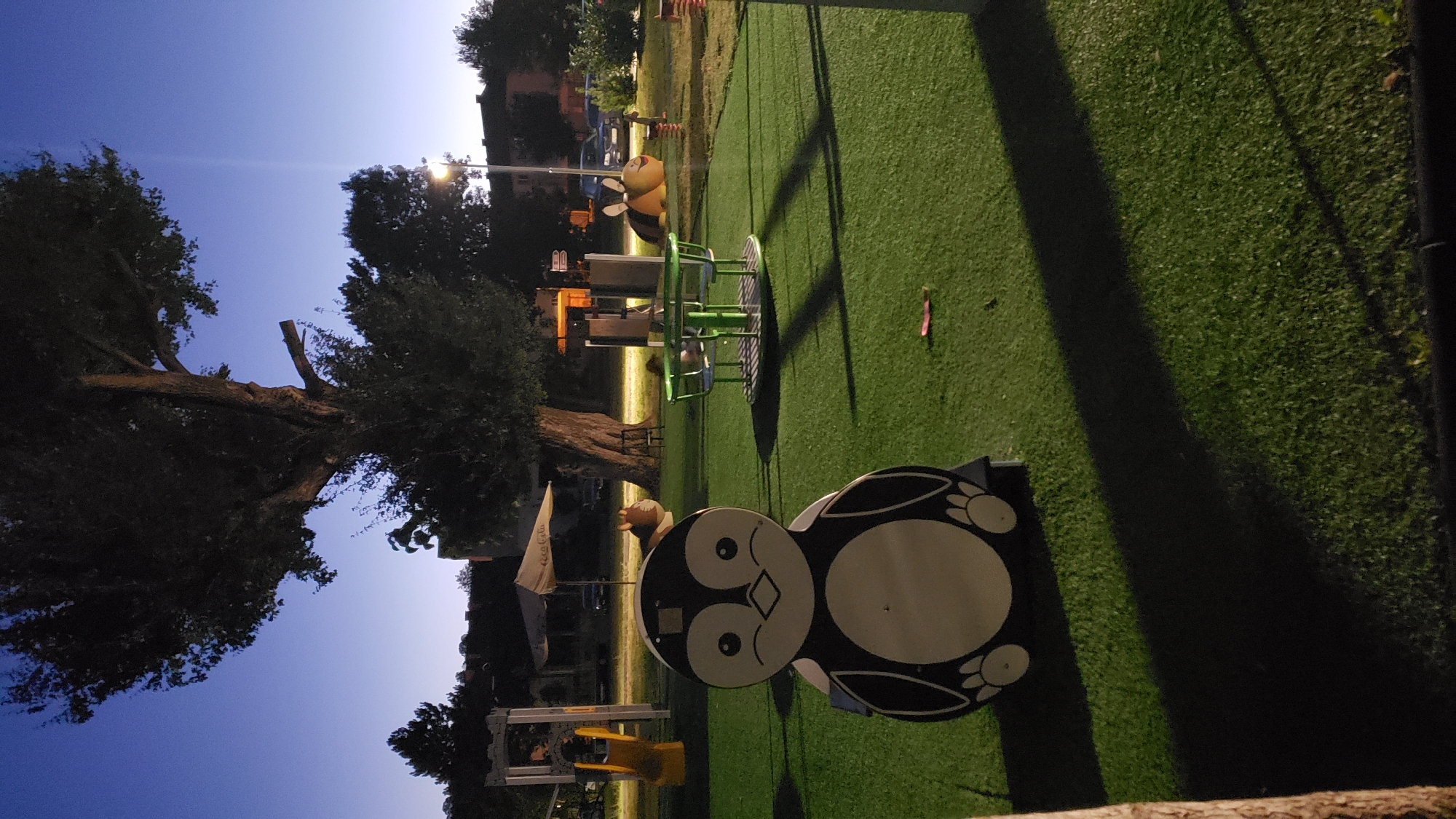 This is a photograph of an outdoor playground at dusk, likely in Prešov, Slovakia. The foreground features a large, two-dimensional penguin-shaped play structure, predominantly black and white, with round eyes and feet. To the left of the penguin is a yellow slide. In the middle ground, a green merry-go-round with a table and chairs is visible. Further back, a large tree dominates the landscape, its dark branches silhouetted against the twilight sky. Beyond the playground, there are glimpses of buildings, parked cars, and streetlights. The ground is covered in artificial turf, and shadows are cast across it. The overall atmosphere is tranquil and serene, with the fading light creating a subdued ambiance.