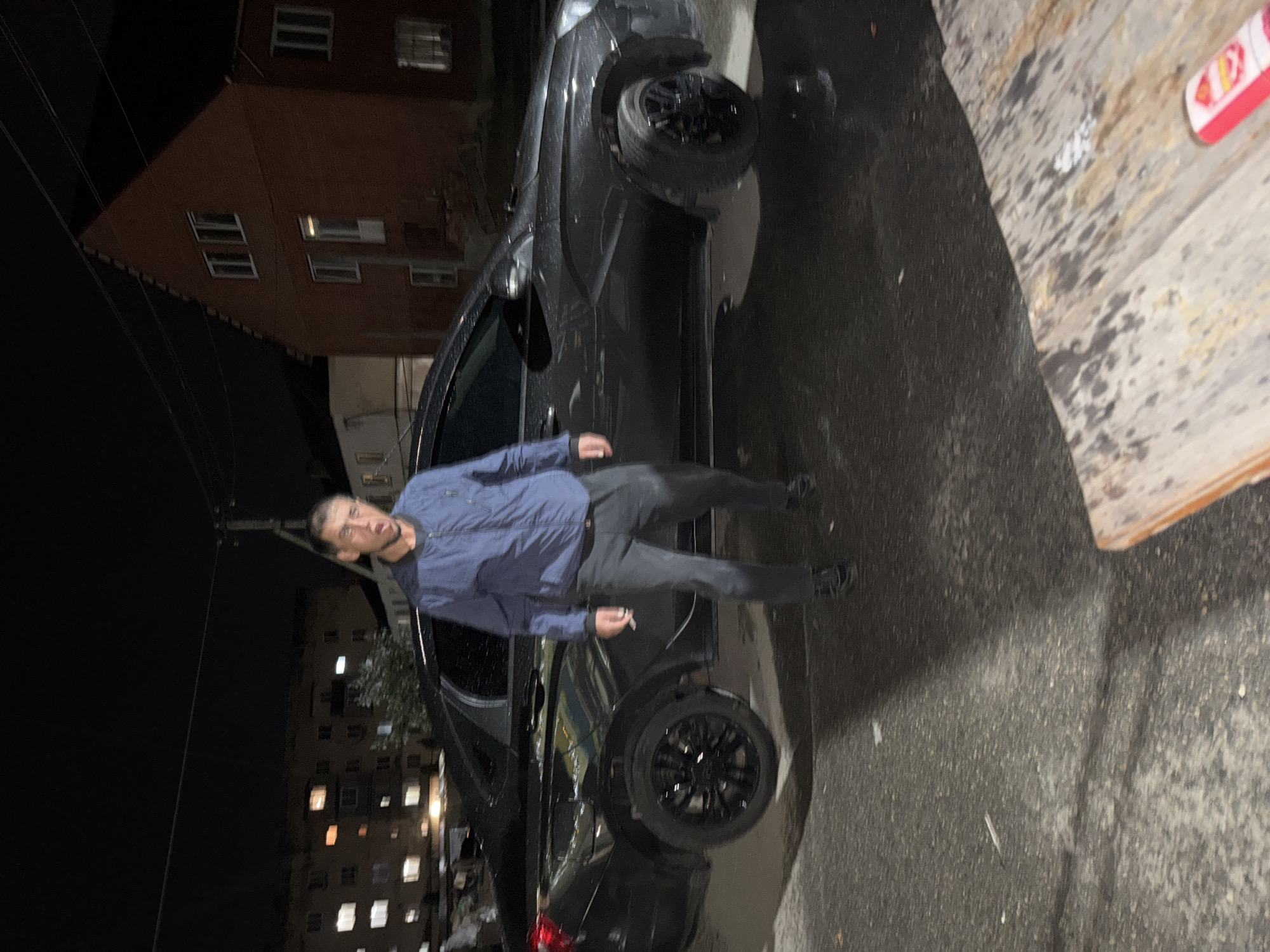 A man stands in front of a black sedan at night. He is wearing a blue jacket and dark pants, with his mouth open as if speaking. The car is parked on a wet asphalt surface, and a wooden table is visible in the foreground. In the background, there are buildings with illuminated windows and a brick structure. The scene appears to be an outdoor urban environment at night.