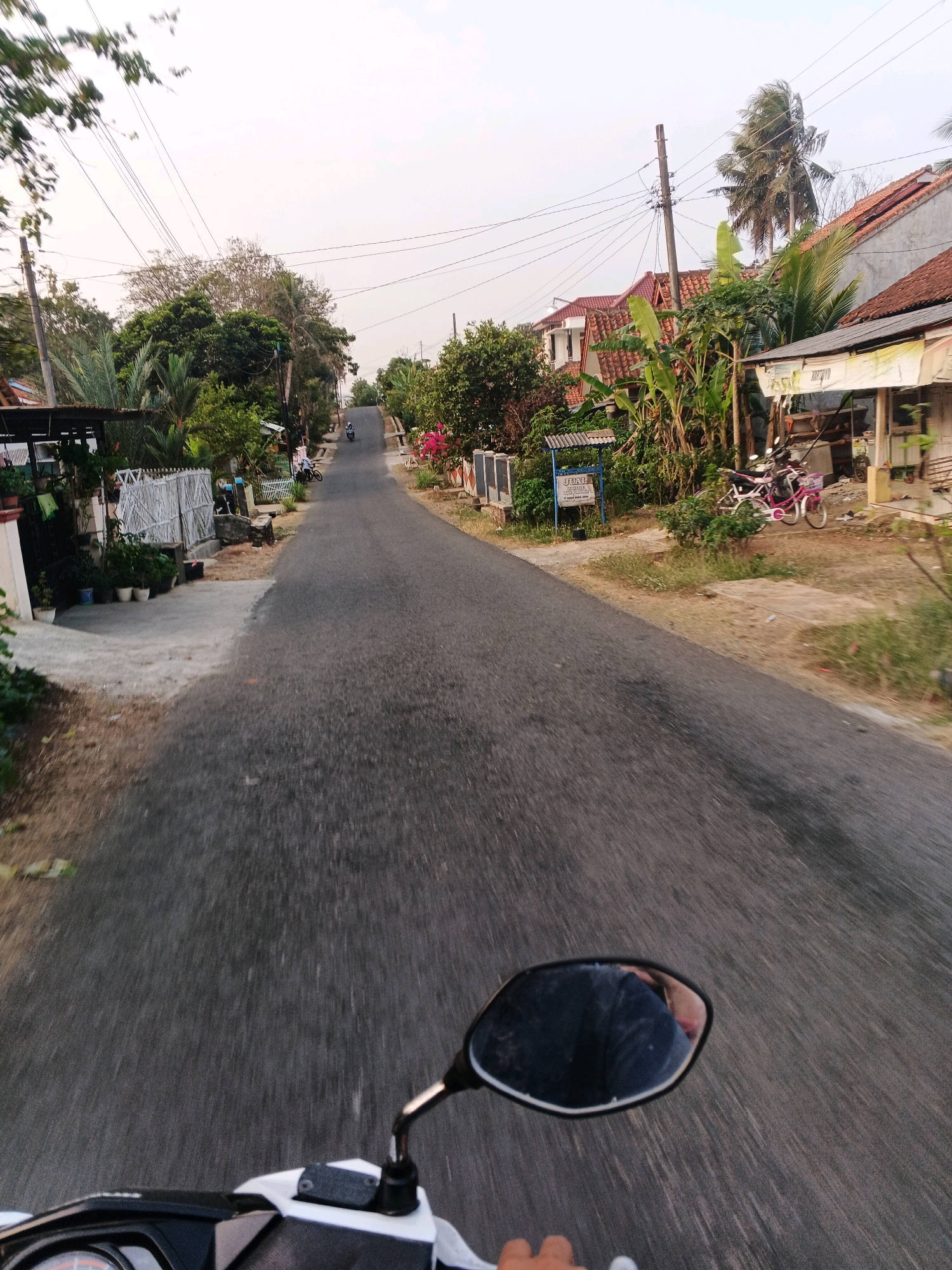 The image depicts a POV shot from a motorcycle traveling down a paved road in Kuripan, Indonesia. The road is lined with houses, lush green vegetation including banana trees and palm trees, and overhead power lines. On the left side of the road, there are houses with white fences and potted plants. On the right side, there are more houses with red tile roofs, and a white stall-like structure. Two bicycles, one pink and one white, are parked near the stall. Further down the road, a person on a motorcycle is visible. The sky is overcast with pale light, suggesting it might be late afternoon or early morning. The reflection in the motorcycle's side mirror shows a person wearing a dark shirt and a glimpse of their face. The image captures a common scene of everyday life in a residential area of Kuripan.