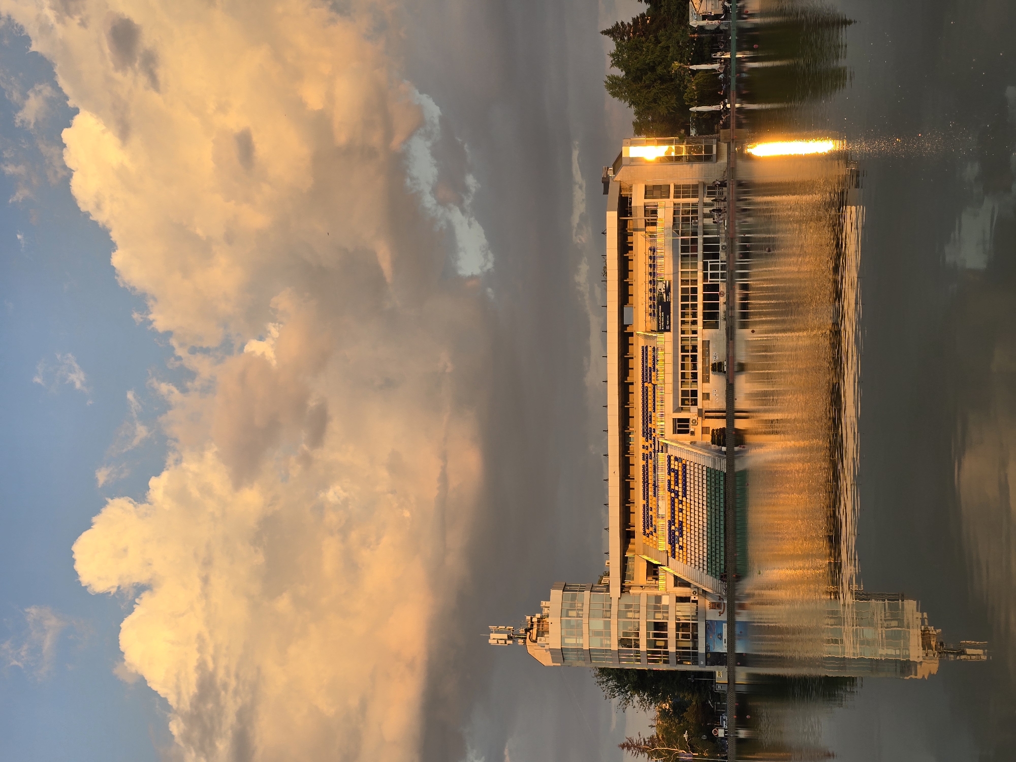 The image shows a building reflected in water. The building is a stadium with seating. The sky is cloudy and there is a sunset. Location: Unknown, Unknown.