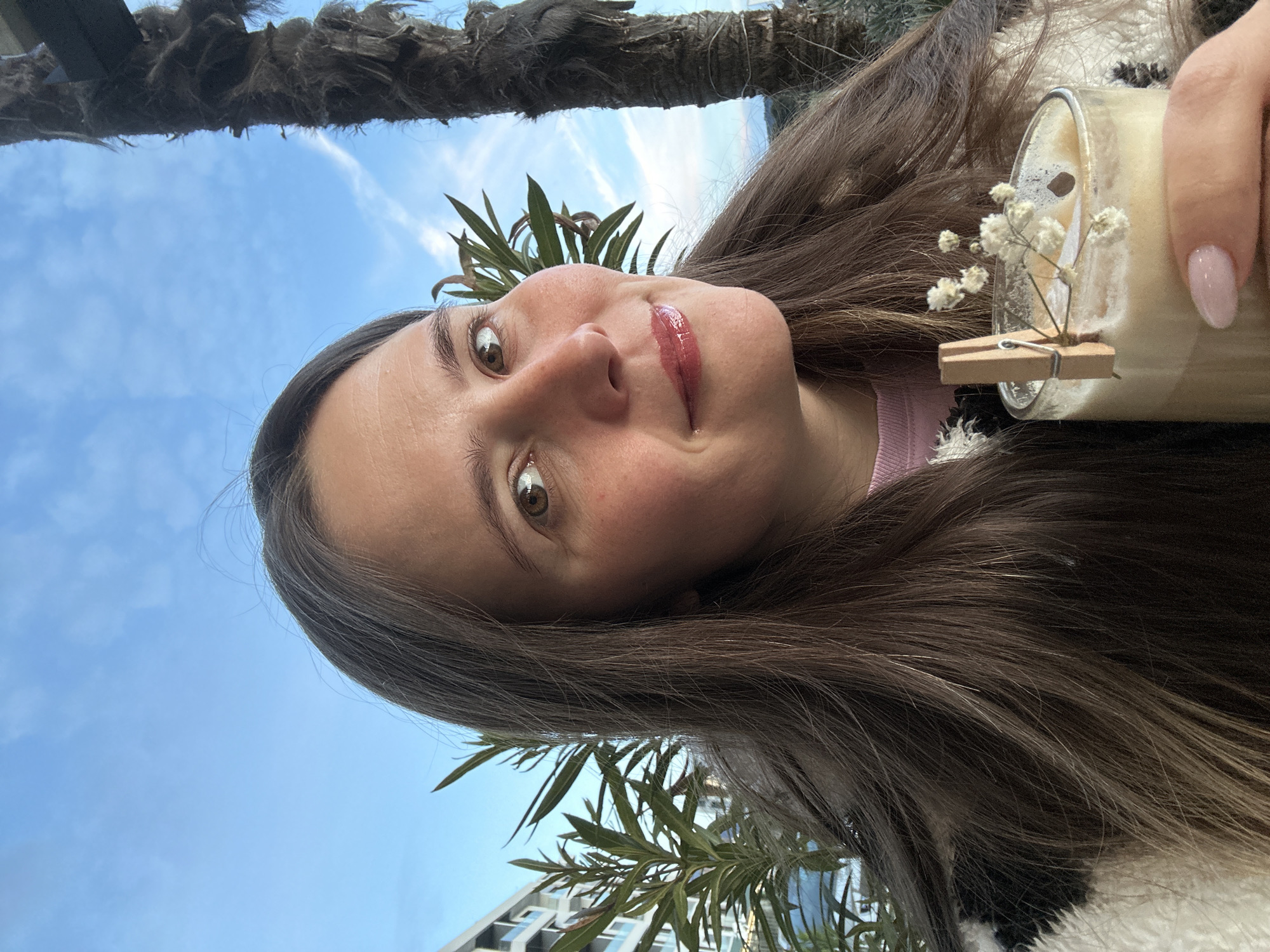 A woman is holding a drink outdoors against a blue sky with scattered clouds. A palm tree is visible above her head. The woman has long brown hair, fair skin, and hazel eyes. She is wearing a pink top and a fuzzy white jacket. She is smiling slightly at the camera. The drink is in a glass with a clothespin clipped to the side, holding small white flowers. In the background, a building with windows and green plants is visible. The image was taken in Šušanj, Montenegro.