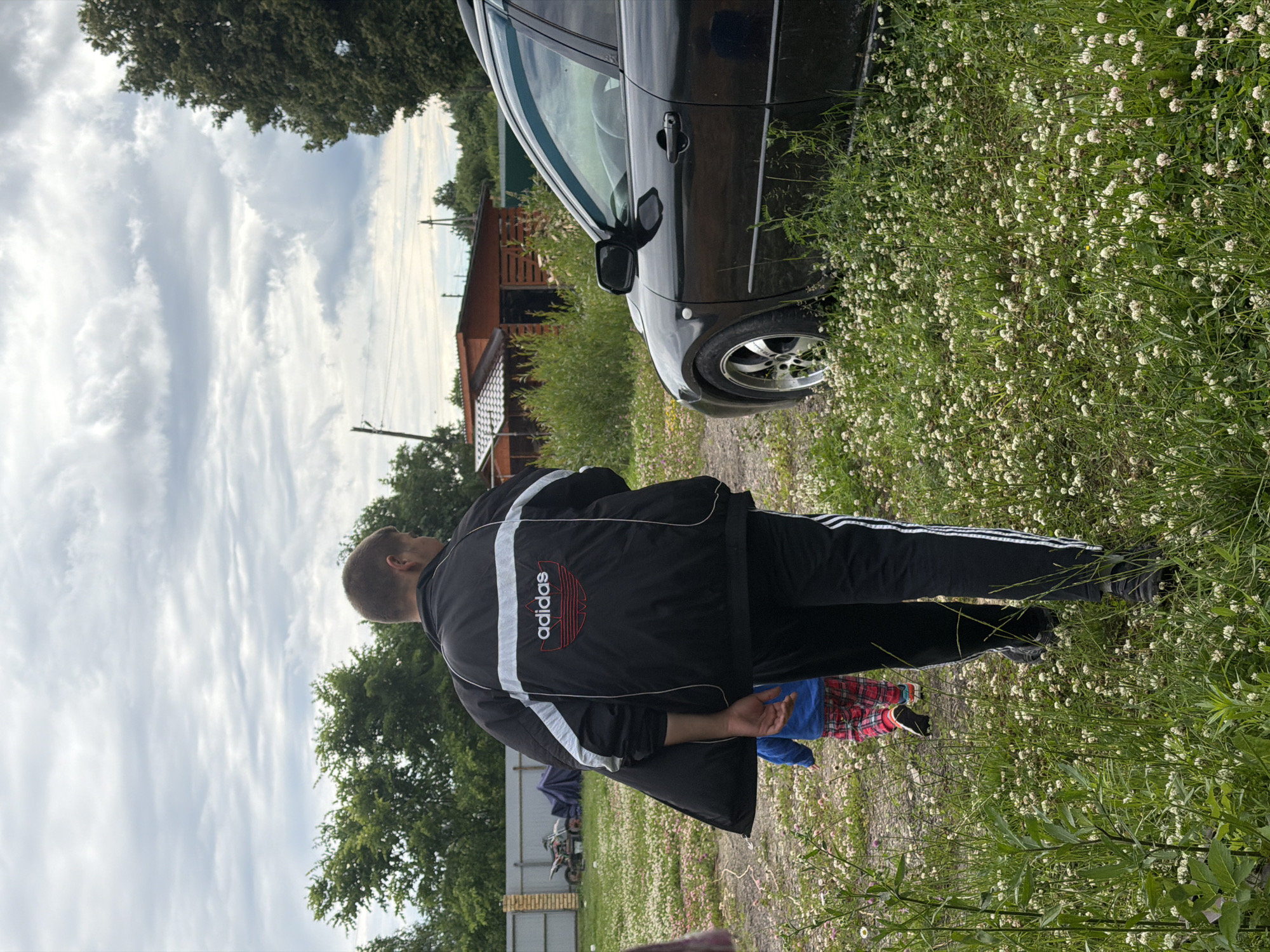 The media file shows a man in an Adidas tracksuit standing next to a black car in a rural setting. The man is viewed from behind, and he appears to be looking towards the left of the frame. The car is parked on a dirt path, with a field of white flowers and green grass bordering it. In the background, there is a wooden structure, possibly a shed or a small house, and trees. The sky is cloudy, suggesting it might be an overcast day. The overall scene appears to be a casual outdoor setting in Andreyevo, Russia, possibly during daytime. The man's attire, the car, and the surrounding environment suggest a relaxed or everyday moment. The Adidas logo is visible on the back of the man's jacket, featuring the iconic three stripes and the brand name.