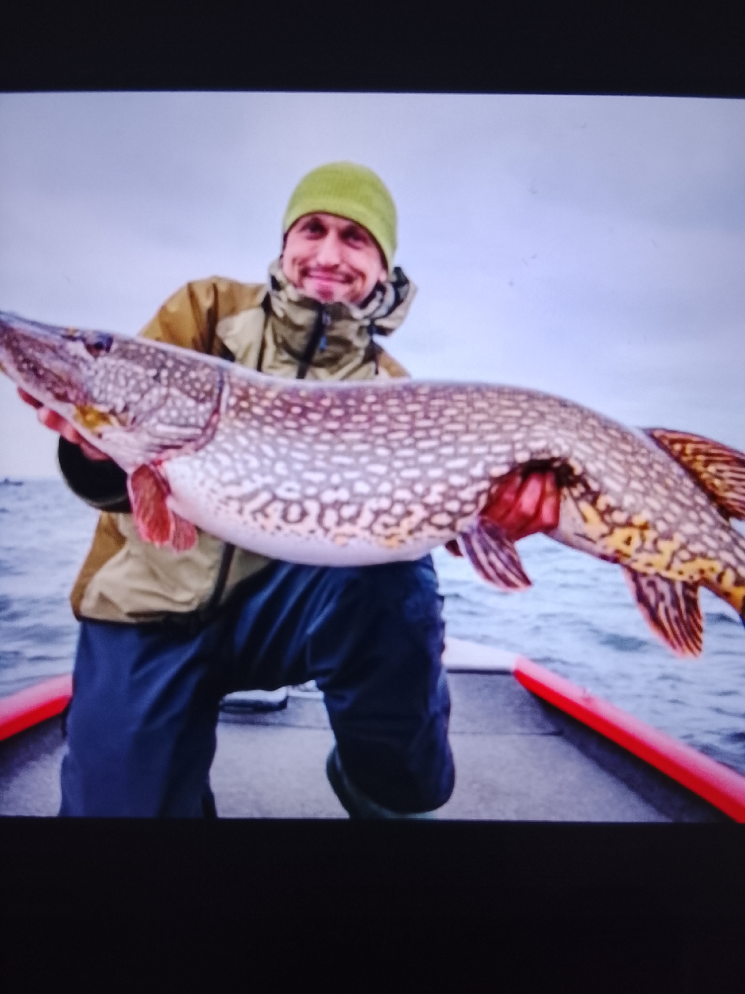 A man wearing a green beanie, a tan jacket, and dark pants is holding a large, speckled fish with red fins. He is in a boat, with the water visible behind him and a red railing in the foreground. The man is smiling and looking at the camera. The sky is overcast.