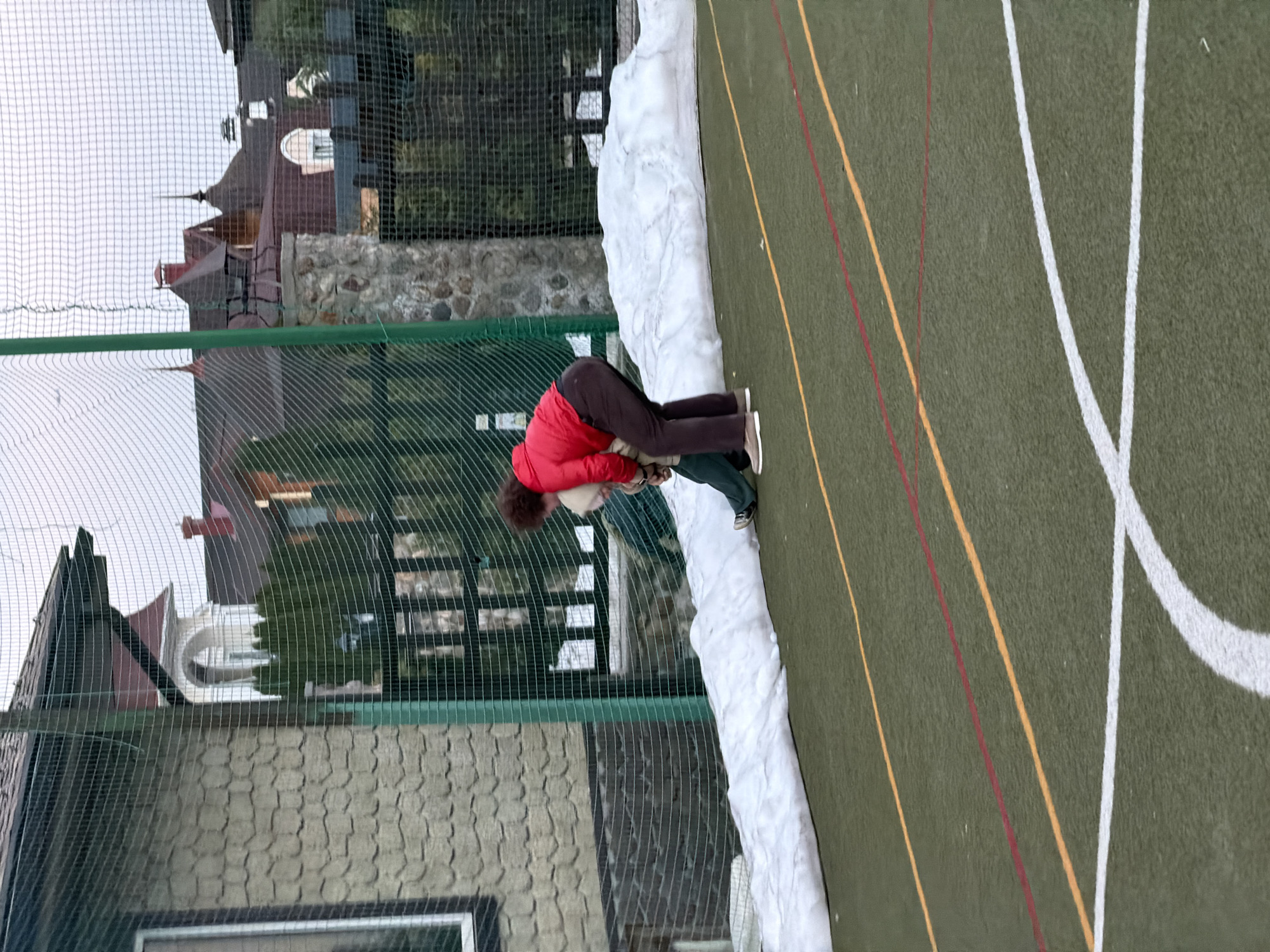 A person wearing a red jacket and brown pants is bending over next to a pile of snow on an outdoor athletic field with green synthetic turf marked with white, yellow, and red lines. Behind the person, there is a green net fence and buildings visible through the fence, suggesting a built-up area. The location is Mishutkino, Russia.