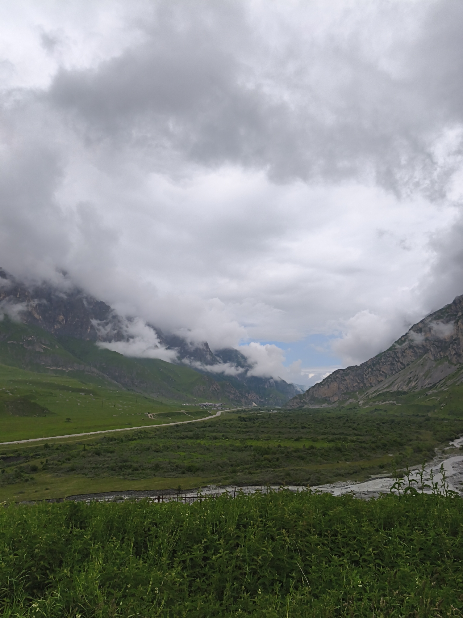The image depicts a wide, verdant mountain valley in Dargavs, Russia, under a heavily overcast sky. In the foreground, lush green grass and vegetation dominate the lower portion of the frame. Beyond this, a wide, grey river flows from right to left, bordered by dense green foliage. A road curves through the middle ground, with a scattering of buildings visible in the distance on the right side, near the base of the mountains. The mountains themselves are imposing and rugged, with steep, rocky slopes partially obscured by dramatic, low-hanging clouds. The overall scene is one of natural grandeur and a somewhat subdued, misty atmosphere typical of a cloudy day in a mountainous region.