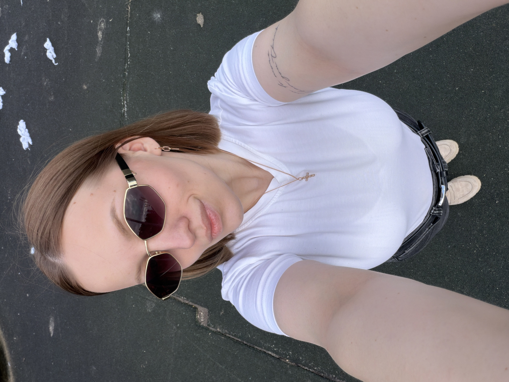 A person, presumably female, is taking a selfie from an upward angle. They are wearing sunglasses, a white t-shirt, dark pants with a belt, and light-colored sneakers. The location appears to be outdoors, with a dark-colored surface visible beneath their feet. The background includes scattered white debris or small objects. There is a tattoo on the person's arm. There is no evidence that the photo was taken in Раздолье, Russia.