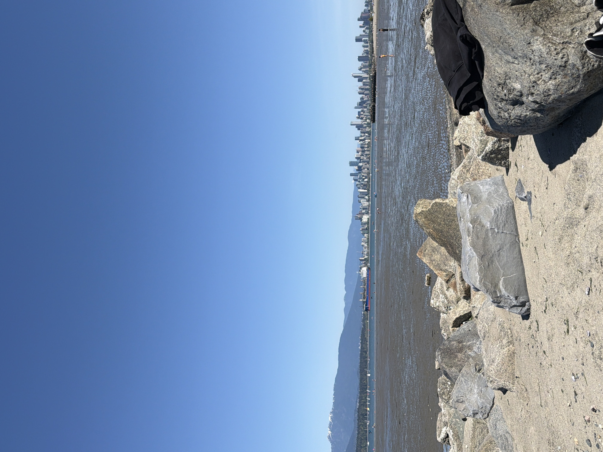 The image shows a bright, clear blue sky over a waterfront scene with a sandy beach and rocky shoreline. A city skyline is visible in the distance, suggesting a coastal urban setting. On the beach, there are silhouettes of people walking along the water. To the right, there are rocks and sand with sparse vegetation. The location is likely a waterfront in Vancouver, Canada, given the mountainous backdrop and urban skyline.
