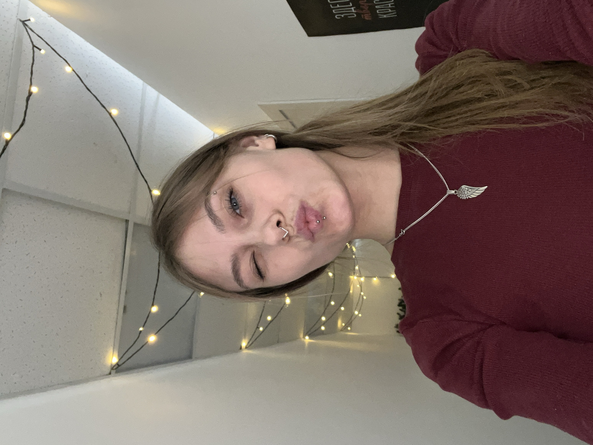 A young, light-skinned woman is posing for a selfie. She has light brown hair, blue eyes, and multiple facial piercings, including a nostril piercing, a lip piercing, and a brow piercing. She is wearing a maroon long-sleeved shirt and a silver necklace with a wing pendant. She is winking and puckering her lips.
In the background, there are strings of fairy lights on a white wall, and a sign with Cyrillic text is partially visible.
It is not possible to confirm the location as Фоки, Russia based on the given image.