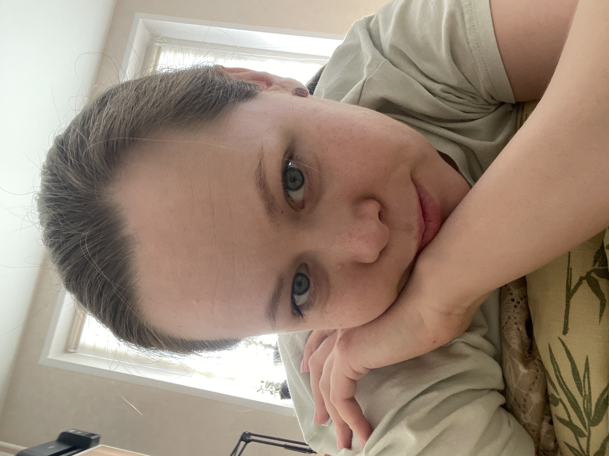 A woman is lying down, resting her head on her hand. She has fair skin, light brown hair, and green eyes. She is wearing a light green t-shirt. The background shows a window with white trim and a wall painted in a light beige color. A desk and various objects are visible in the lower left corner. The location is potentially a home in the city of Chaikovsky, Russia, as requested, but there is no forensic information present in the image that could independently confirm the location. The picture is taken at an angle. The woman appears to be looking directly at the camera with a soft expression.