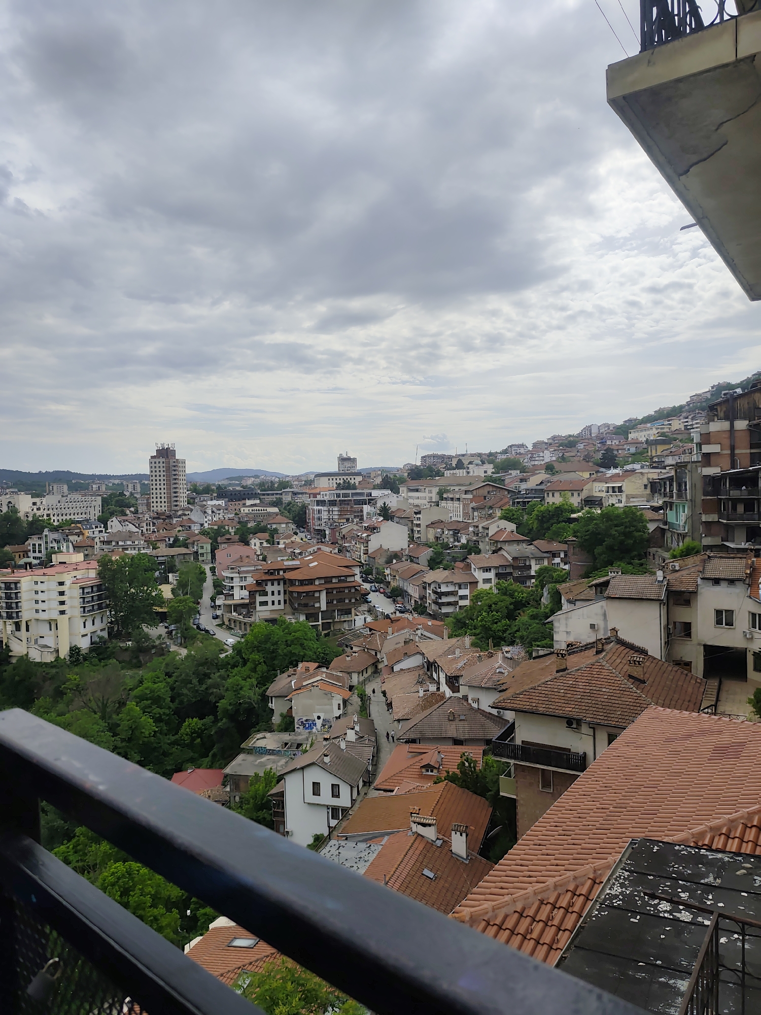 An elevated view of the city of Veliko Tarnovo, Bulgaria under a cloudy sky. Buildings with red-tiled roofs dominate the foreground and midground, interspersed with green trees. The city stretches into the distance, with taller buildings and mountains visible on the horizon. A portion of a balcony with a black railing is visible in the bottom left of the image. A concrete overhanding roof with metal railing is visible in the upper right.