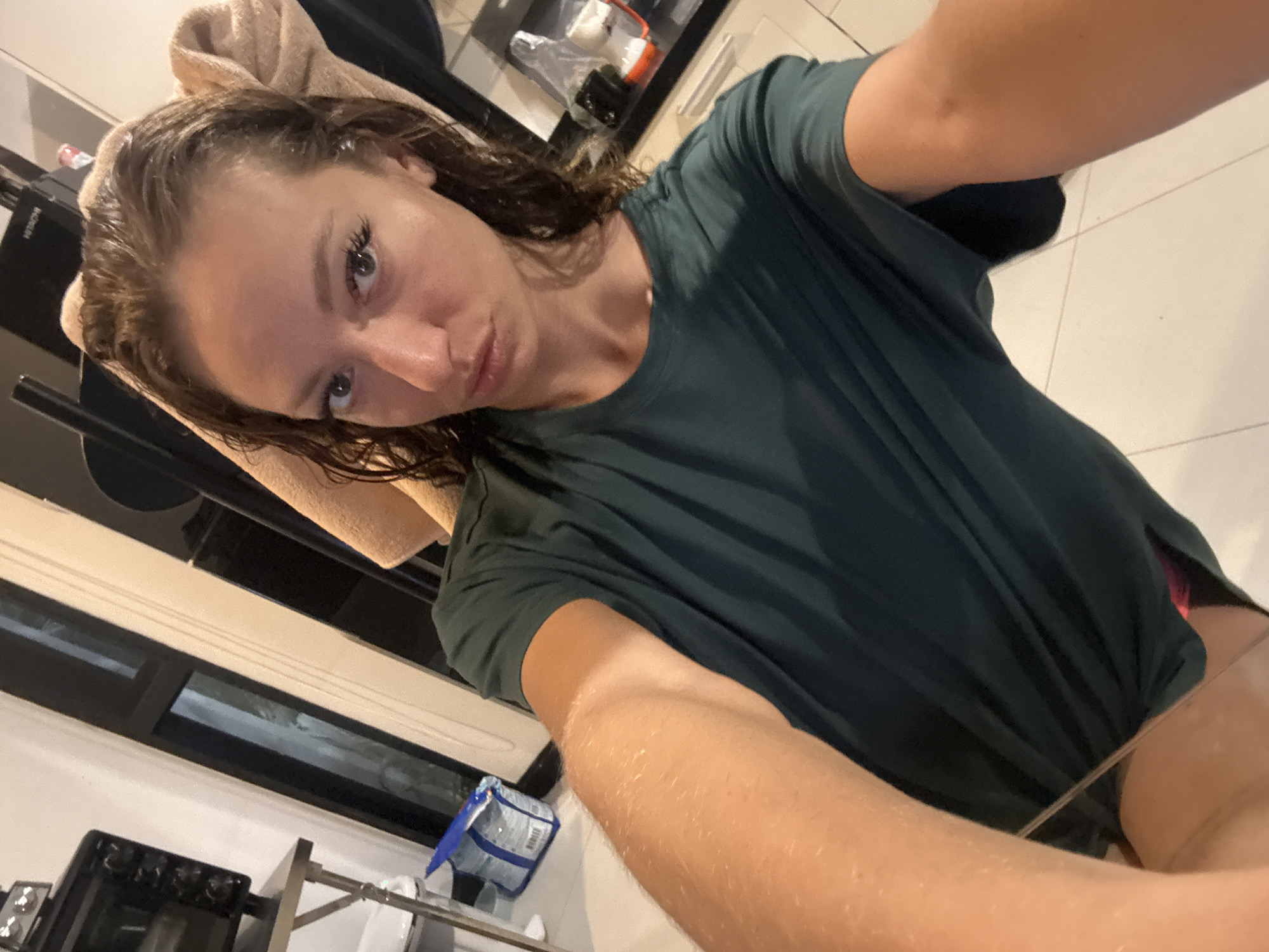 A young woman takes a selfie in an indoor setting, possibly a kitchen or bathroom in Baan Chaweng, Thailand. She has wet hair wrapped in a beige towel and is wearing a dark green t-shirt. The background shows a window, kitchen appliances, and tiled flooring. A package is visible below. The woman is holding the camera at an angle and has a slight pout.