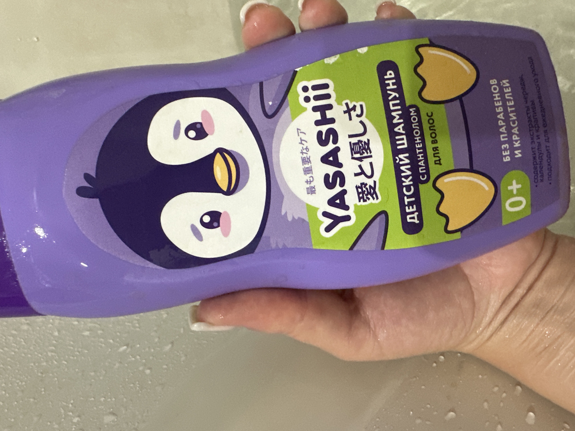 A person's hand, with manicured fingernails, holds a purple bottle of "YASASHii" children's shampoo. The shampoo bottle features a cartoon penguin graphic and text in Russian and Japanese. The background is a wet, light-colored surface, possibly a shower wall. There is no identifiable information in the image to determine its location in Novokuznetsk, Russia.