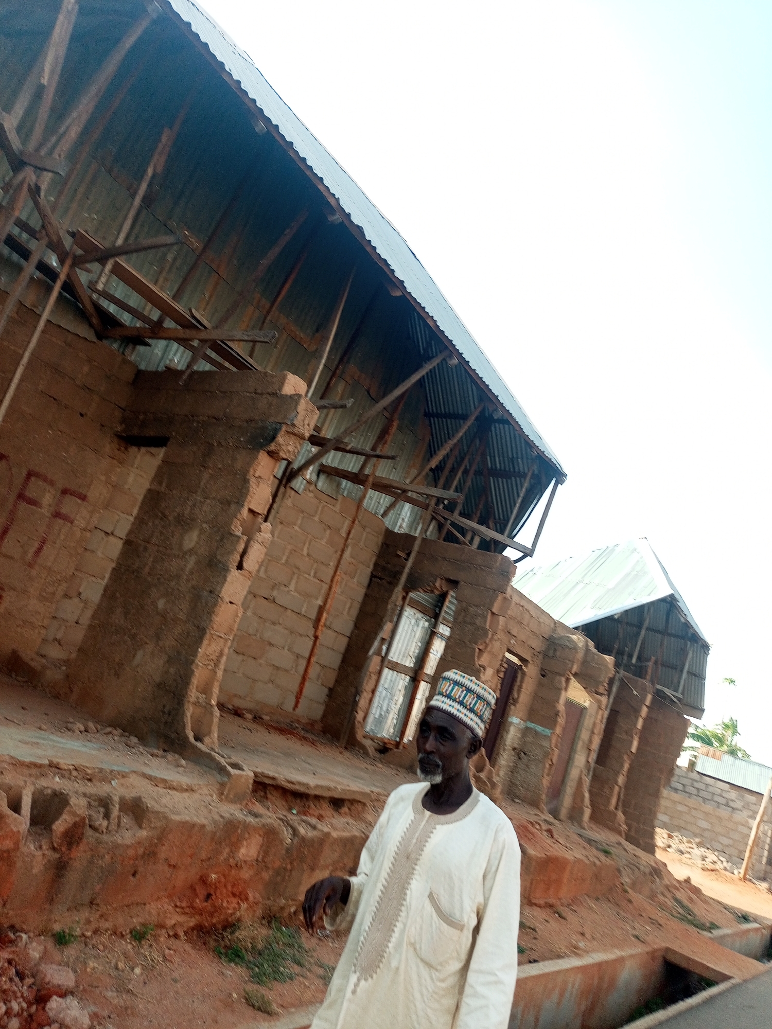 An elderly man stands in front of a dilapidated structure in Tumfure, Nigeria. He is wearing a white robe with embroidered details and a patterned cap. The building behind him is partially destroyed, with brick walls and a corrugated metal roof supported by exposed wooden beams. The surrounding area appears to be an impoverished neighborhood.