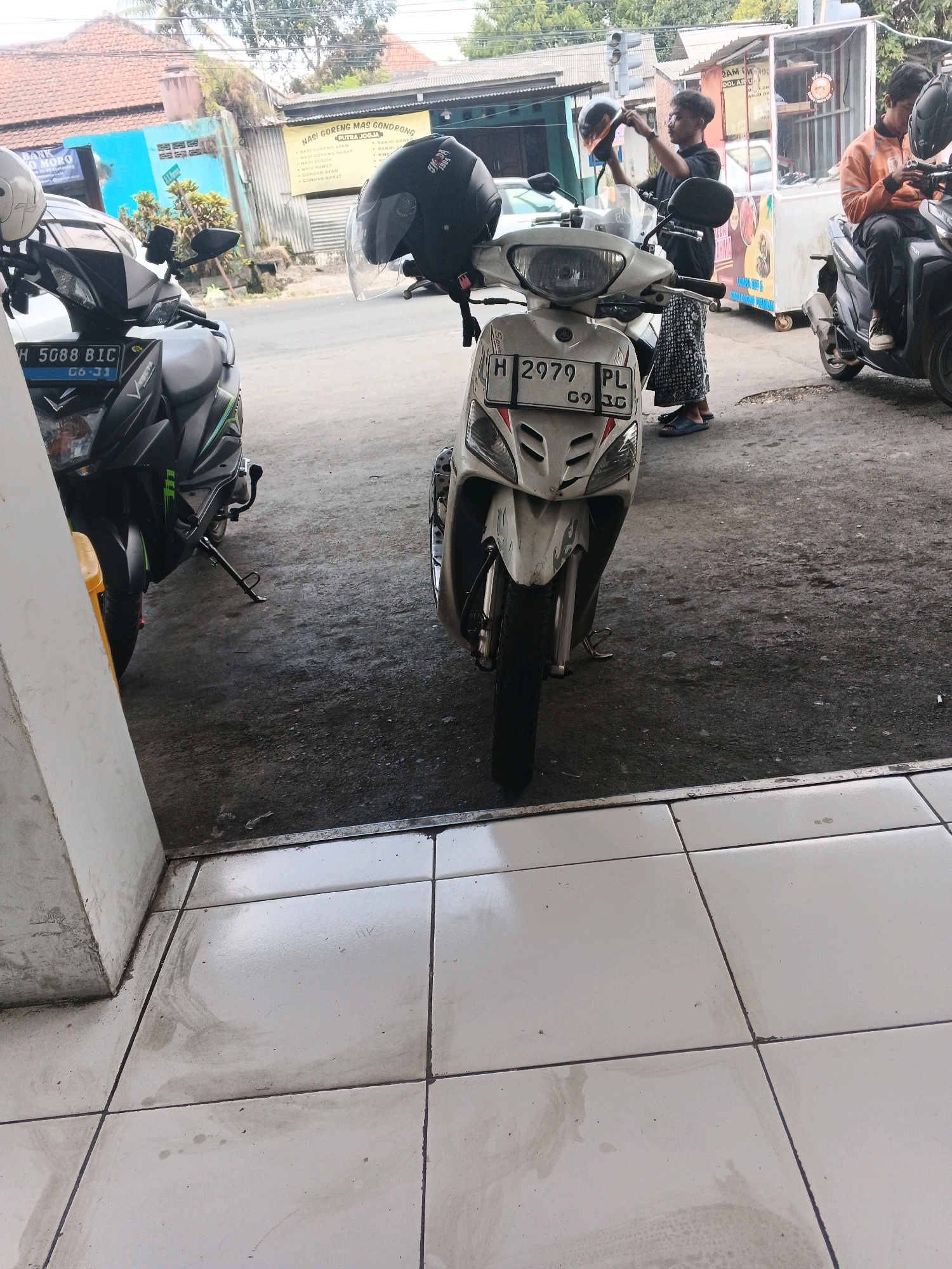 In Genuk Barat, Indonesia, two motorcycles are parked outside a food stall. A white motorcycle with a black helmet on its handlebars is in the foreground. A black motorcycle with a rider wearing an orange shirt is to the right. Behind the motorcycles, a food stall with a glass display case and menus is visible. The ground is a mix of asphalt and tiled pavement.
