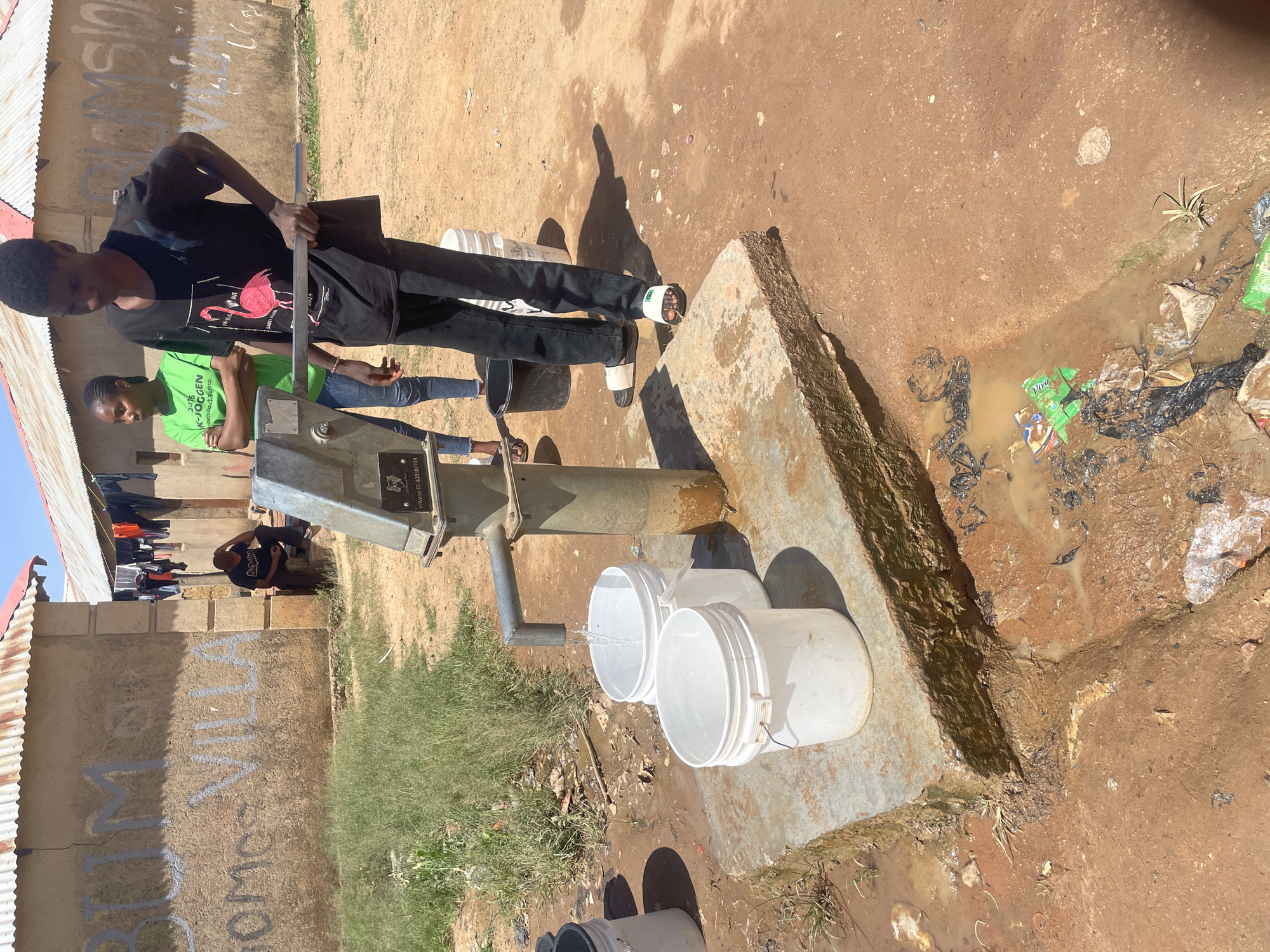 Two people are operating a water pump in Durimi, Nigeria. One person is pumping water into white buckets. The other person is standing next to the pump. The area around the pump is muddy and littered with trash. A building with graffiti is in the background.