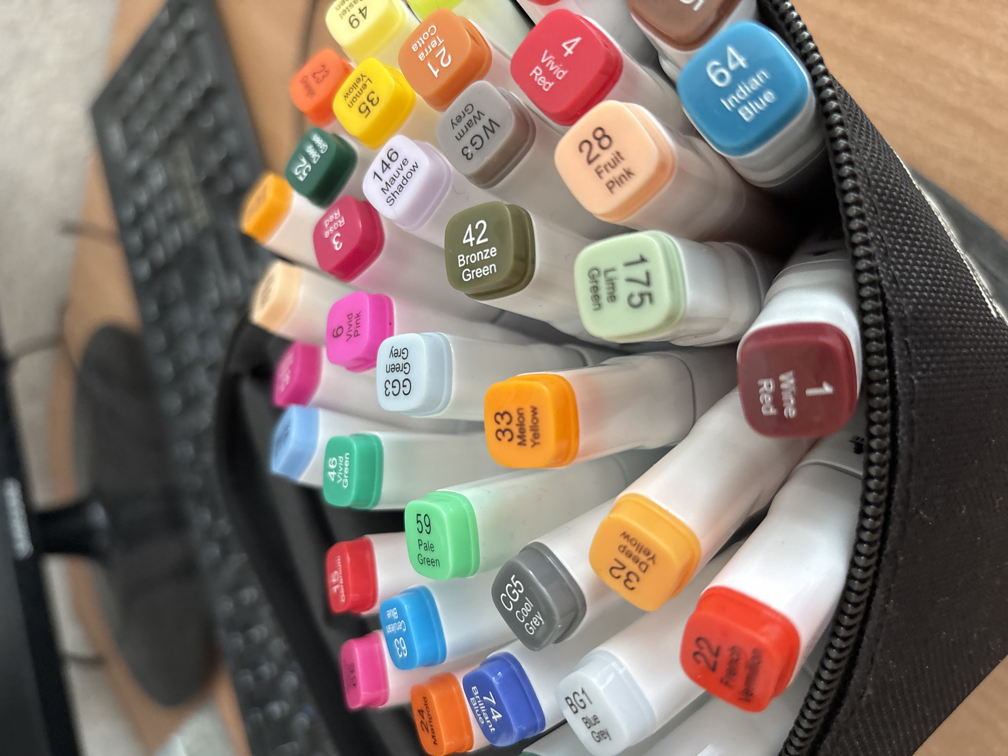 An overhead, eye-level shot shows a black zippered pouch filled with markers. The markers are arranged in a tightly packed, overlapping manner. The caps of the markers display different colors and numbers, with some color names also visible. In the blurred background, a black computer keyboard and monitor are visible, suggesting an indoor setting. The lighting is natural, casting soft shadows on the markers. The image composition focuses on the details of the markers, their colors and labels. There is no indication of the city of Sochi, Russia, or any specific geographical location.