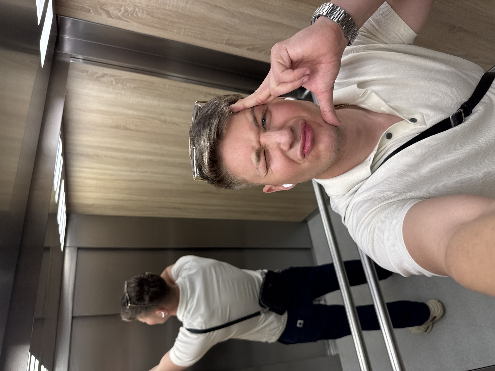 The image shows a man taking a selfie in an elevator. The man has fair skin, short blonde hair, and is wearing a white collared shirt, a silver watch, and sunglasses resting on his head. He is winking and making a silly face. The elevator has wood-paneled walls and a metal handrail. Reflected in the elevator's mirrored wall is the man's lower body, wearing dark jeans, white shoes, and a black bag across his body. The location appears to be inside a building in a generic elevator; without further context, it is impossible to determine if it is located in Plovdiv, Bulgaria.