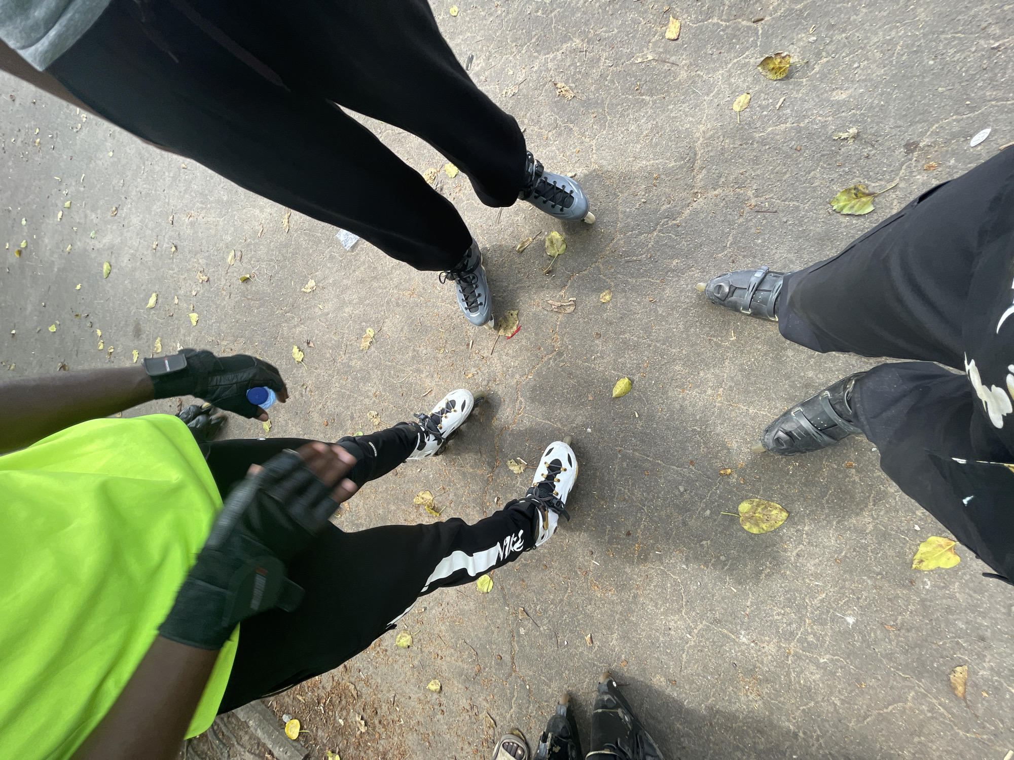 The image shows a group of people wearing rollerblades standing on a paved outdoor surface. The ground is cracked and appears to be concrete or asphalt, with scattered fallen leaves indicating an outdoor, possibly park-like setting. One individual is wearing a bright neon green shirt and black pants with white accents, and is also wearing black fingerless gloves. Another individual is wearing black pants and grey rollerblades. A third individual is wearing black pants and white rollerblades with black laces and accents. A fourth person's legs and rollerblades are visible in the foreground, wearing black pants and black rollerblades. One person is holding a blue water bottle. The overall scene suggests a casual gathering or activity involving rollerblading. The location is specified as Alagbede, Nigeria.
